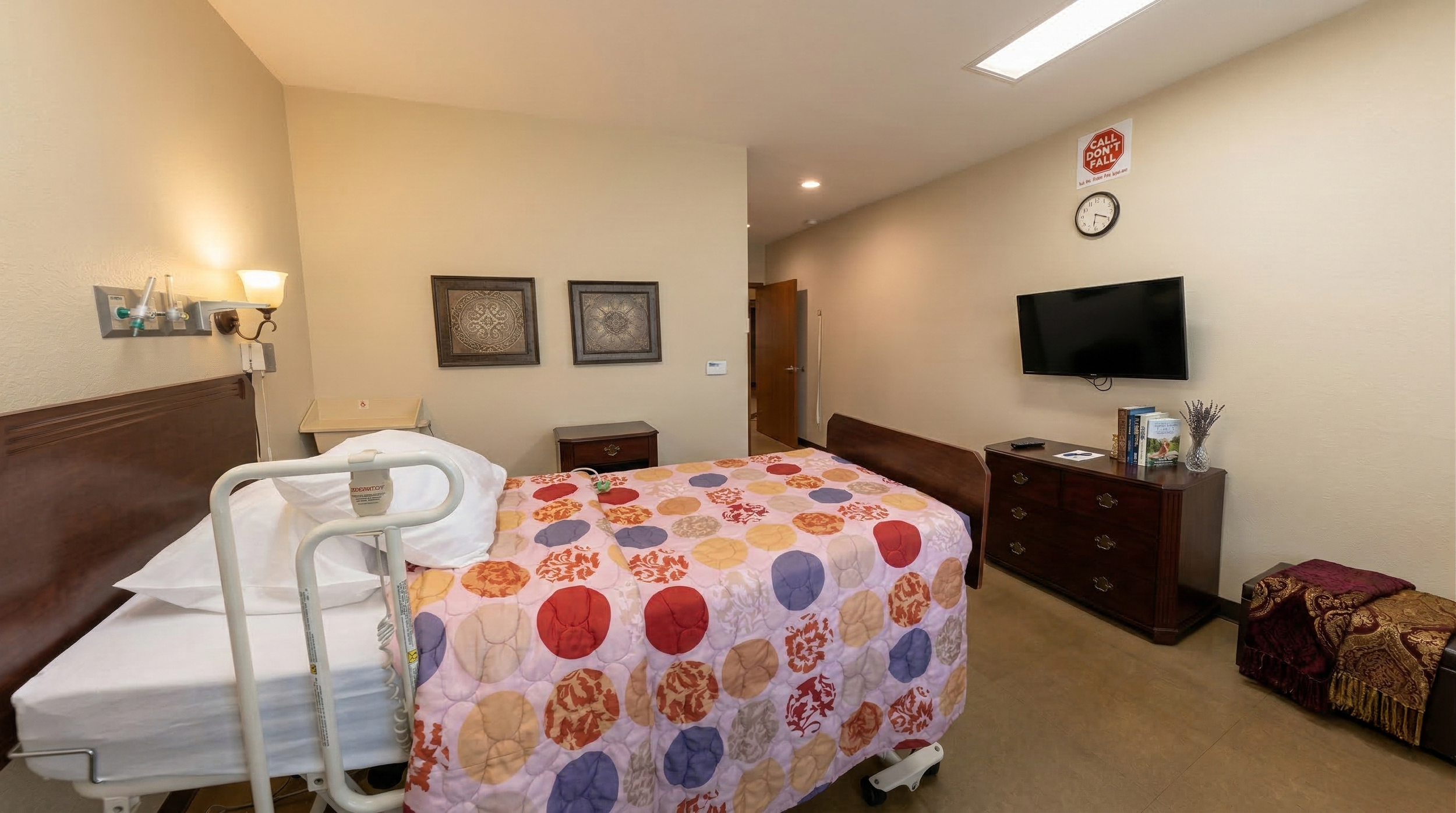 Nursing home room with a bed covered by a colorful quilt, medical equipment attached to the headboard, a wooden nightstand, a dresser with a TV above, framed pictures on the wall, a clock, and a sign that says 'Call Don't Fall'.