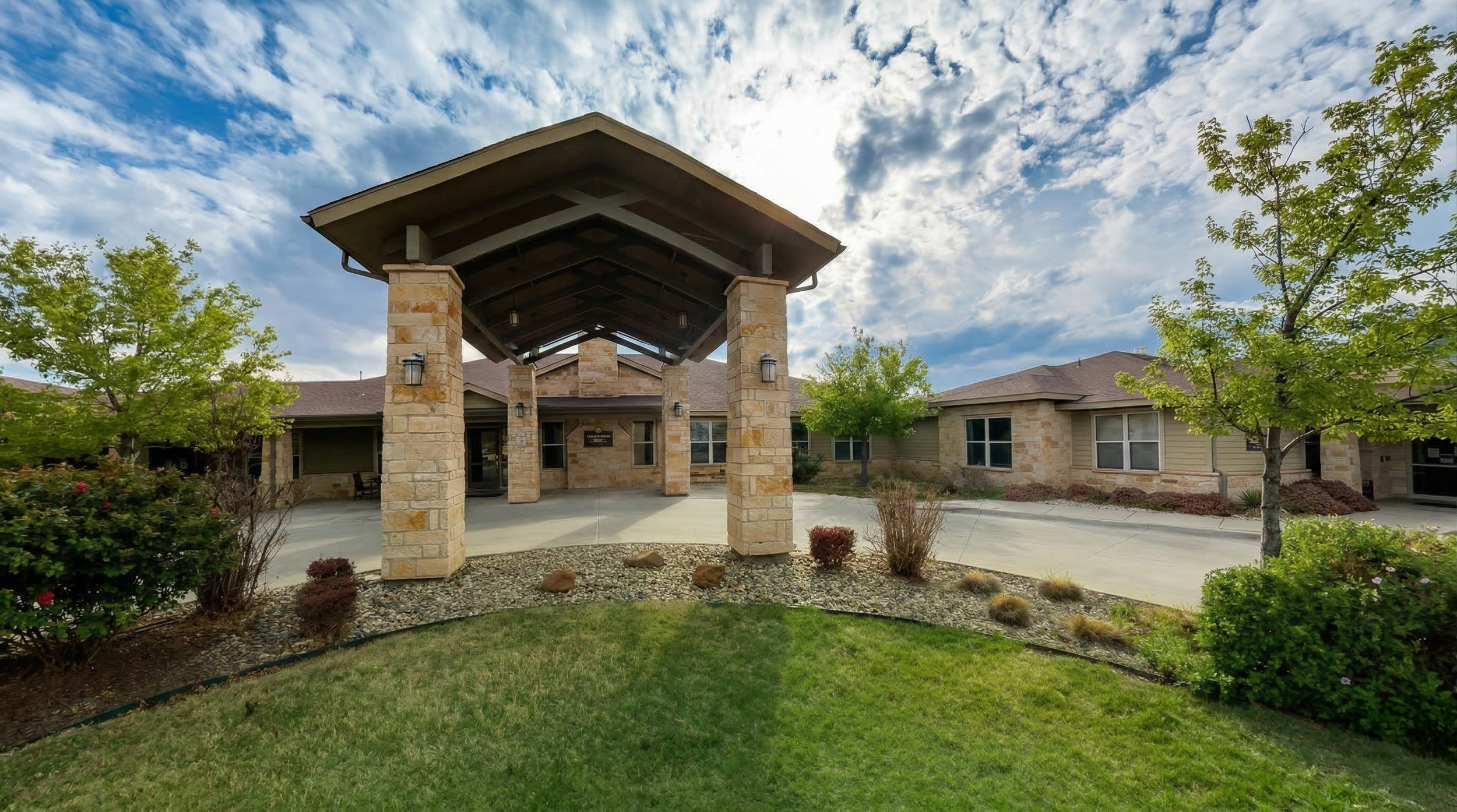 Exterior view of a modern building with a stone entrance, green grass, trees, and a partly cloudy sky.