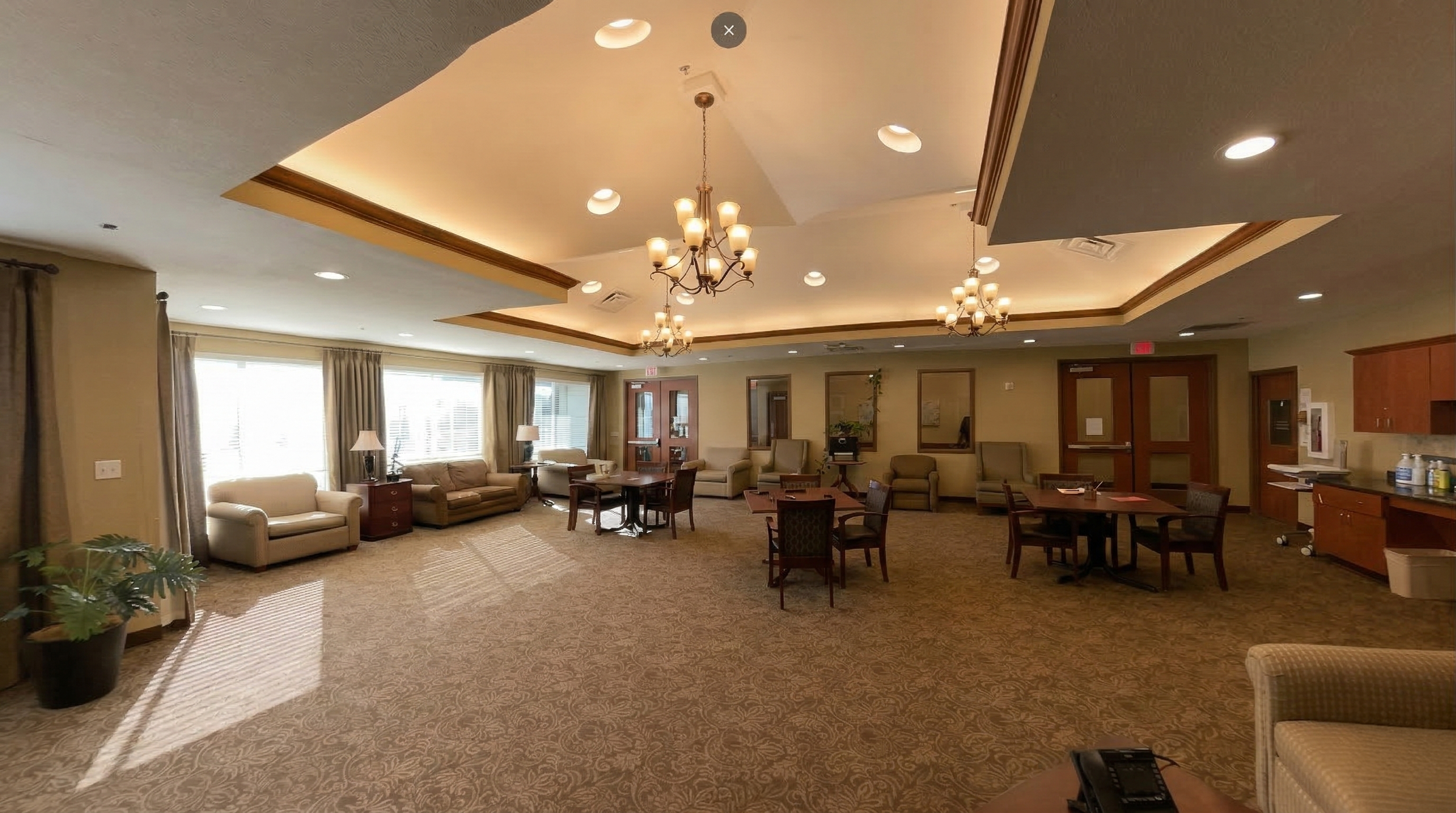 Spacious activity room and event center with beige sofas, wooden chairs, round tables, lamps, windows with curtains, and chandeliers, resembling a hotel lounge or community center.