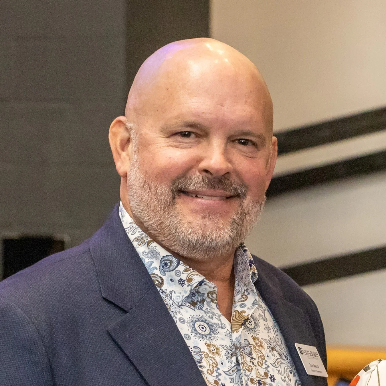 Dan Marick wearing a paisley patterned shirt and a navy blazer, standing indoors with a blurred background.