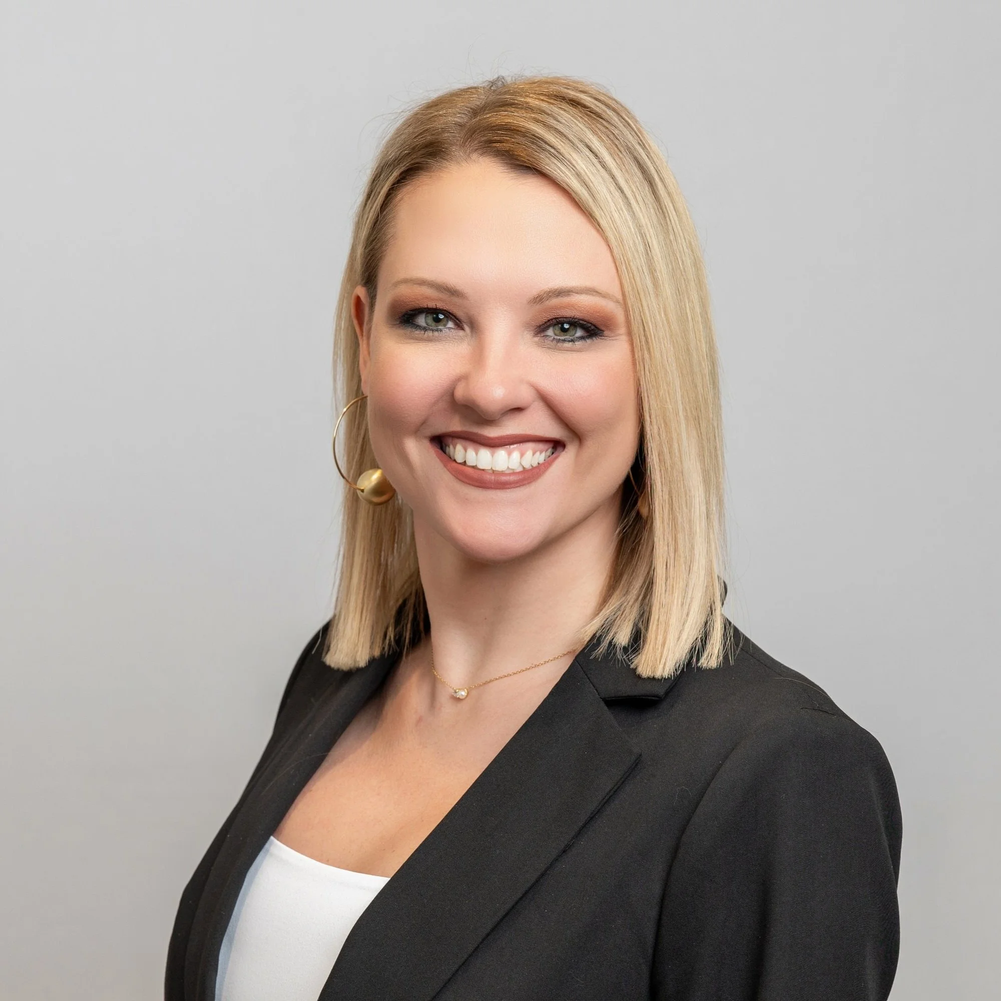 Heather Burchett wearing a black blazer, white top, gold hoop earring, and a delicate gold necklace, against a plain light grey background.