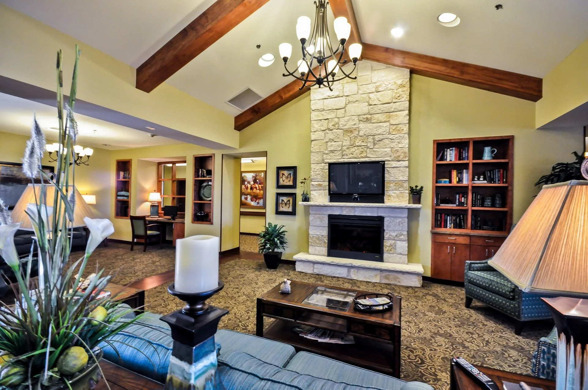 Spacious living room with high vaulted ceiling, exposed wooden beams, and a large stone fireplace with a flat-screen TV mounted above. Contains a blue upholstered sofa, side tables with lamps, a coffee table, bookshelves, and decorative plants. A chandelier hangs from the ceiling.