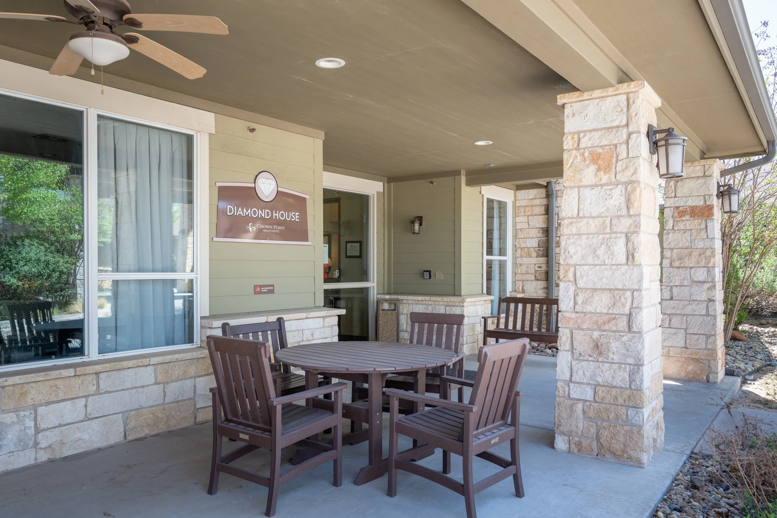 Outdoor patio area with a round wooden table and four chairs, stone pillars, and a sign that reads "Diamond House" on a light green building.