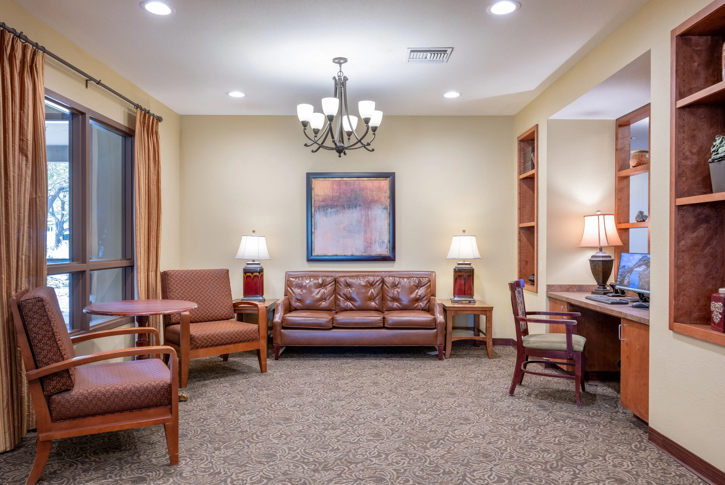 Living room with beige walls, wooden furniture, brown leather sofa, two matching lamps, armchairs, a window with curtains, a desk with a computer, and framed artwork on the wall.