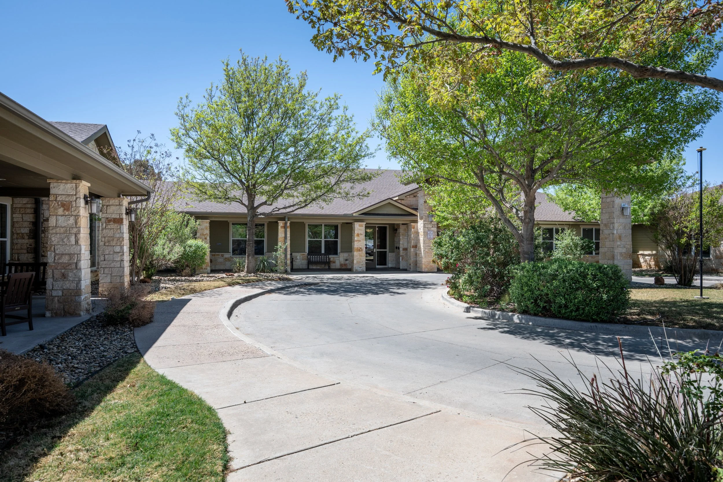 A nursing facility entrance with a circular driveway, multiple mature trees providing shade, and well-maintained landscaped bushes and grass. The buildings are made of stone and siding with covered porch areas.