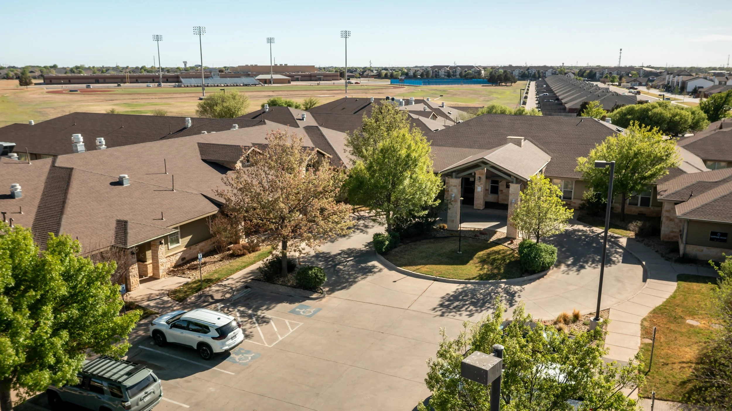 Luxury senior care community with a parking lot, surrounded by trees, and a sports field in the background.