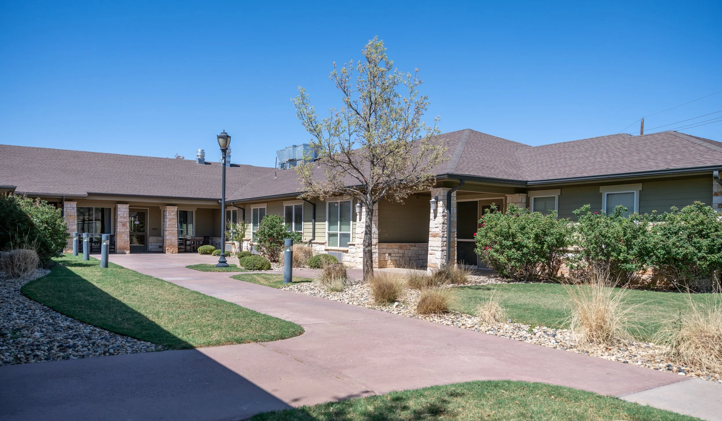 Exterior view of a senior living complex with a central walkway, landscaped with grass, shrubs, and trees. The building has green siding, stone accents, and a brown shingled roof, with a blue sky in the background.