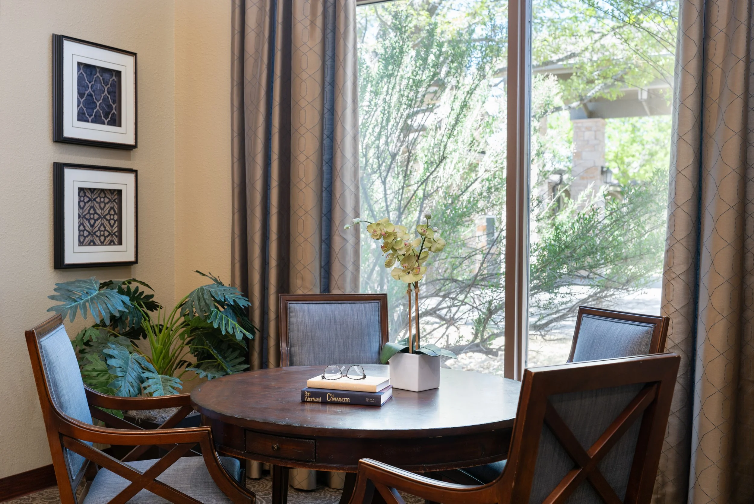 A round wooden dining table with four chairs, a pair of glasses, a stack of books, a small potted plant, and a large window with curtains showing outdoor greenery.
