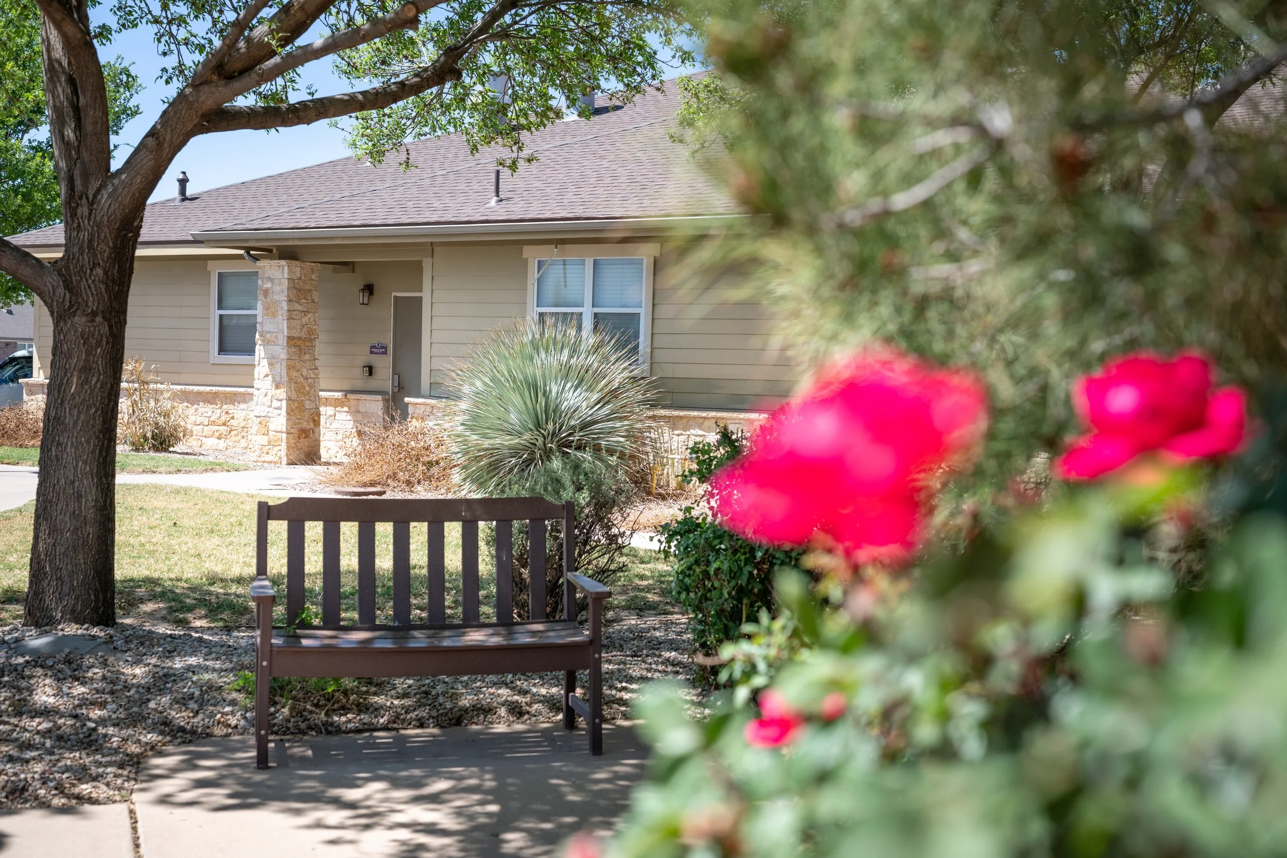 Post-acute care facility front yard with a wooden bench, trees, flowering bushes, and a house with beige siding and stone accents.