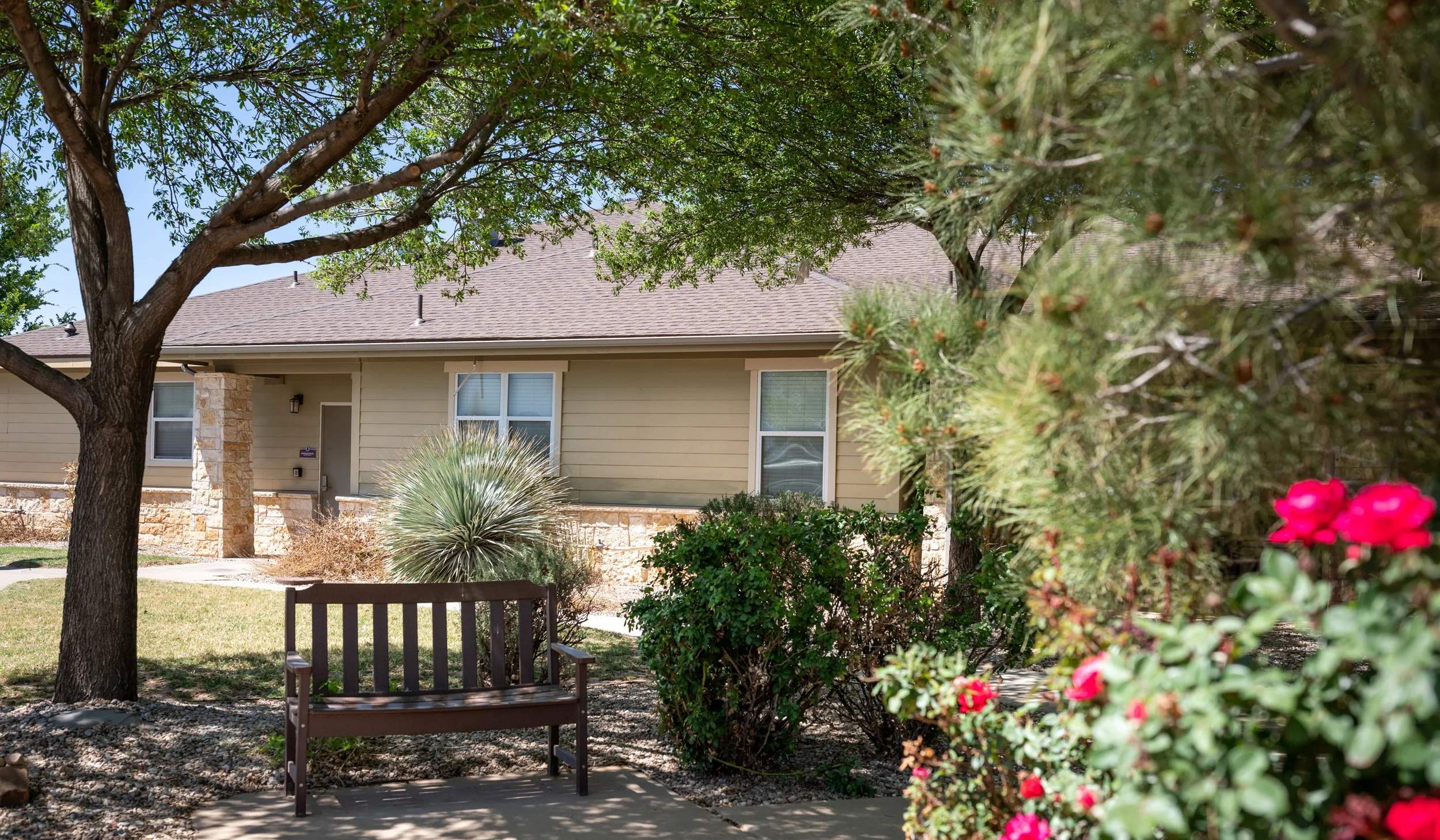 A residential house with beige siding, stone accents, and two visible windows, surrounded by landscaped greenery including trees, bushes, and flowering plants. A wooden bench is placed on a small concrete slab near the sidewalk. The scene is sunny, w