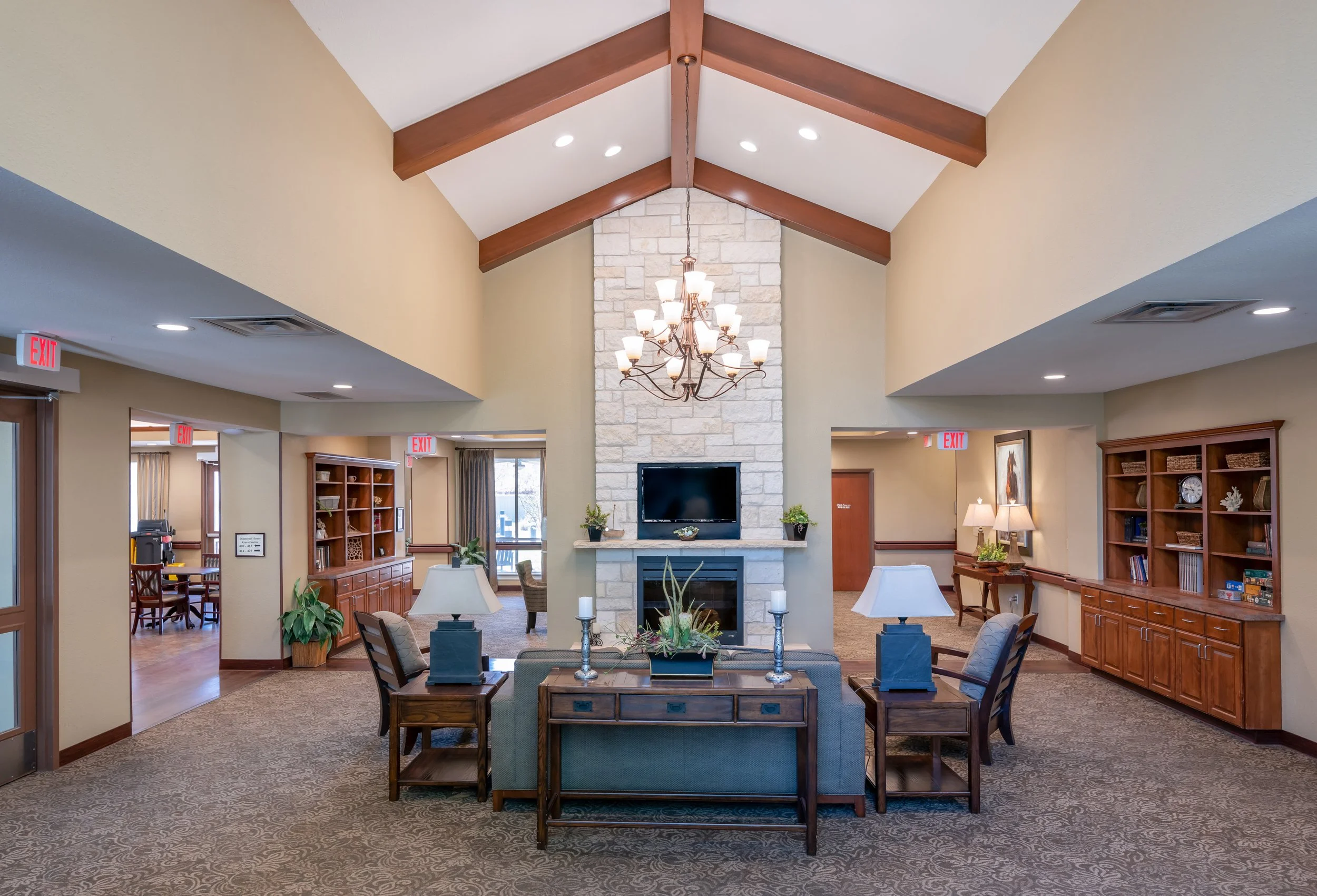 Spacious hotel-like nursing home lobby with a high ceiling, stone fireplace, chandelier, and comfortable seating area with tables and lamps.