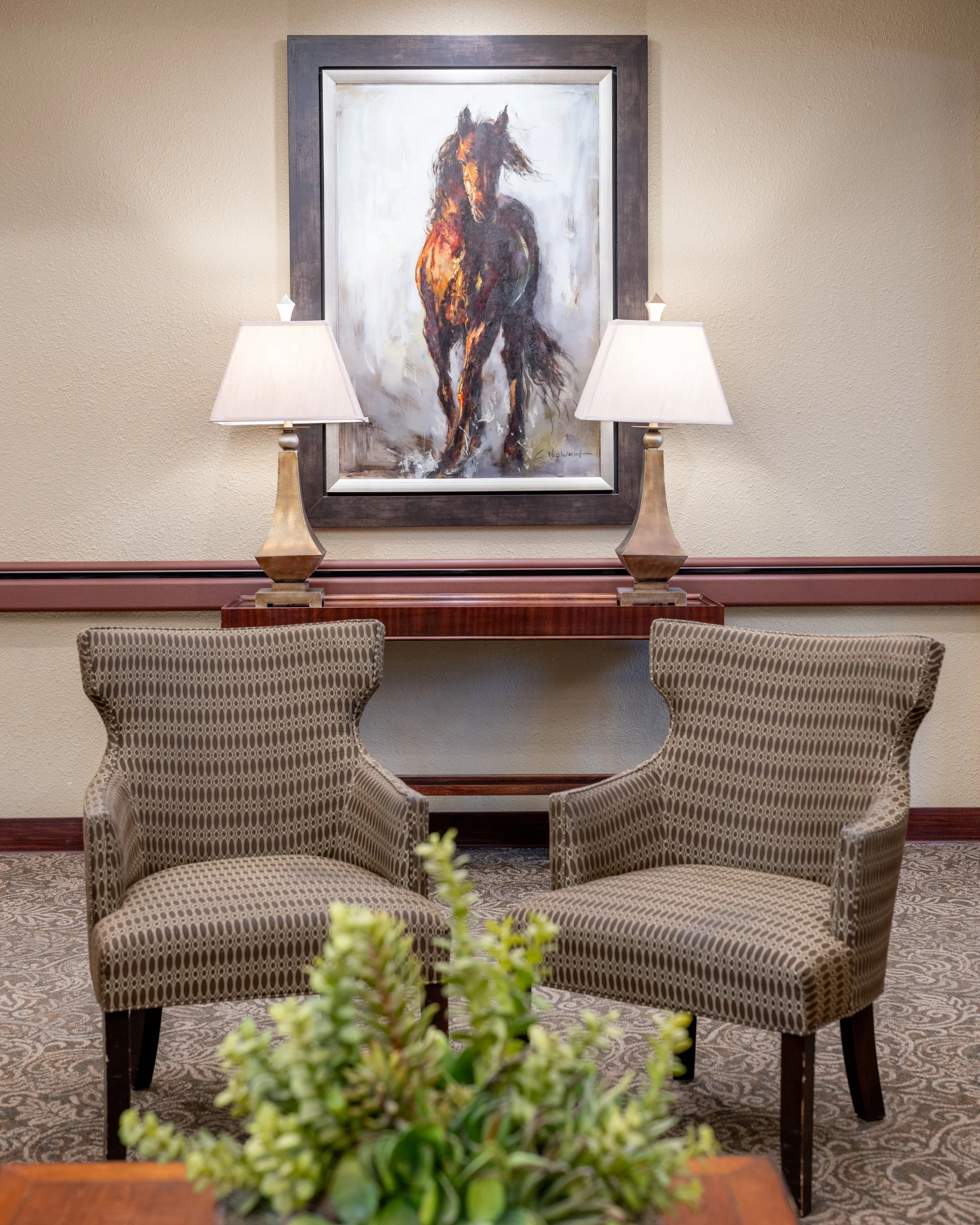 Interior of SNF lobby area with two patterned armchairs, a wooden table, two table lamps, a painting of a running horse, and a potted plant in the foreground.