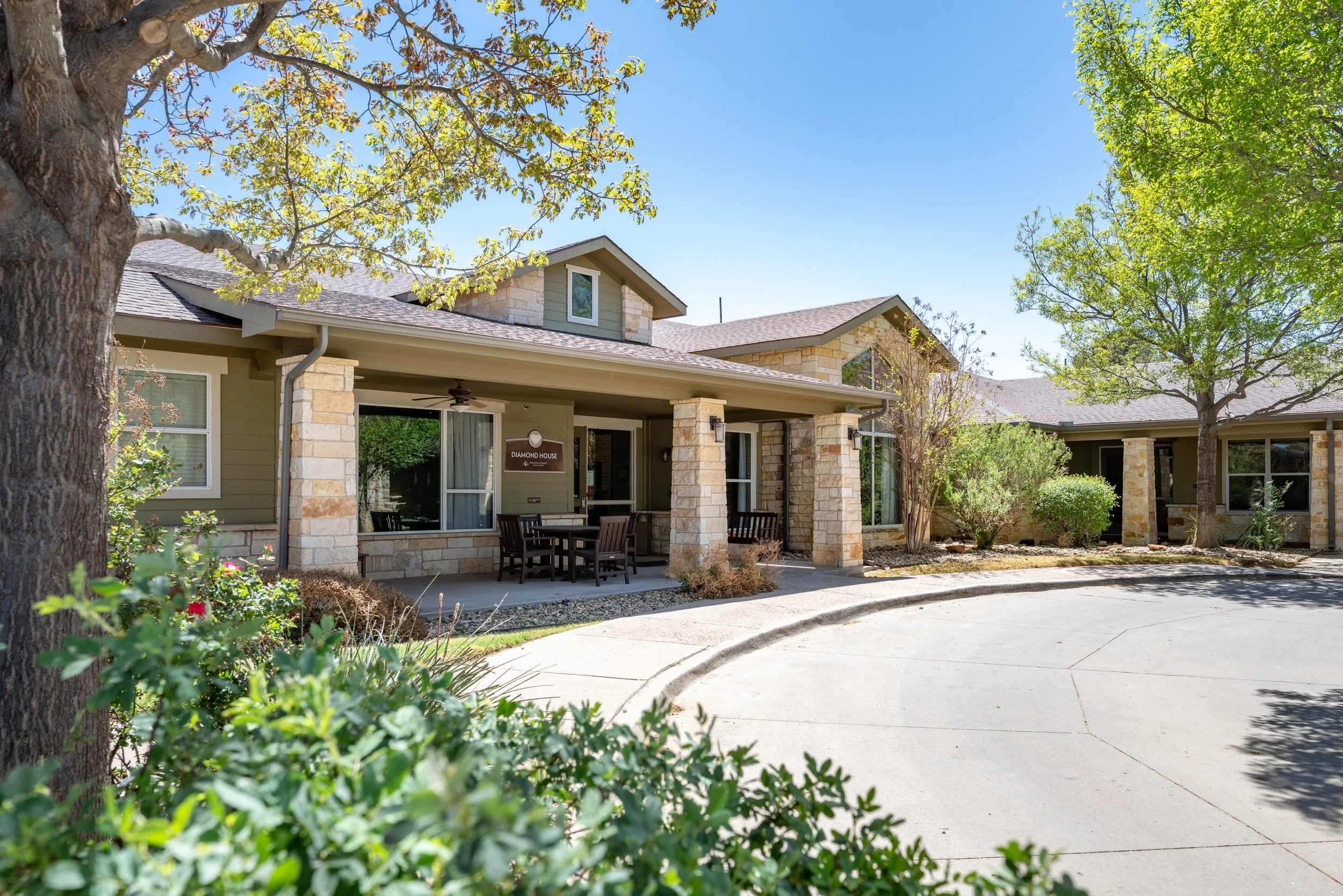 Front of a Lubbock, Texas senior care facility with trees and a curved driveway, featuring stone and siding exterior, large windows, and a porch with outdoor chairs.