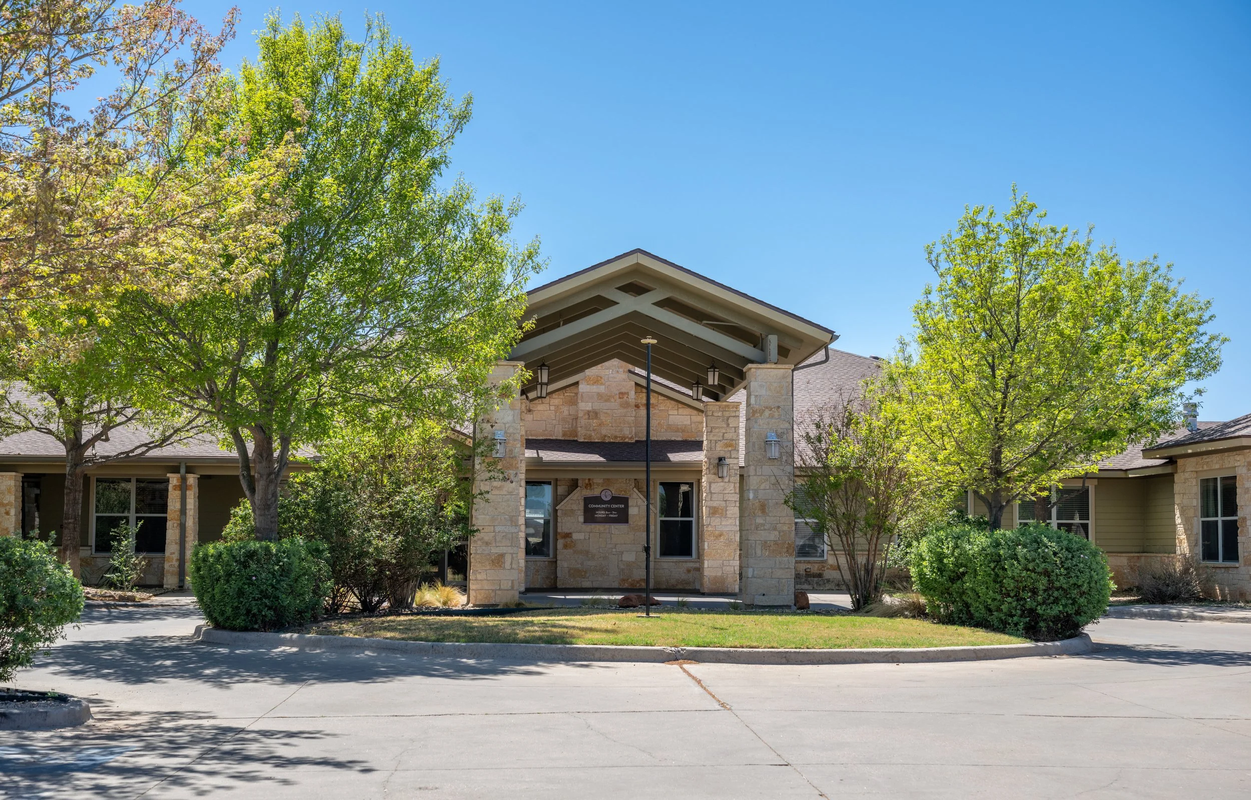 Front view of a senior community building with stone walls, surrounded by trees and bushes, on a clear sunny day.