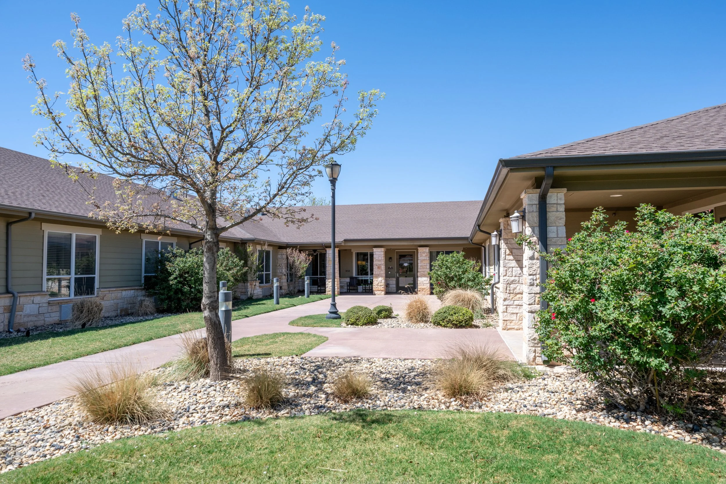 Lubbock, Texas skilled nursing facility courtyard with trees, bushes, a sidewalk, and a lamp post under a clear blue sky.