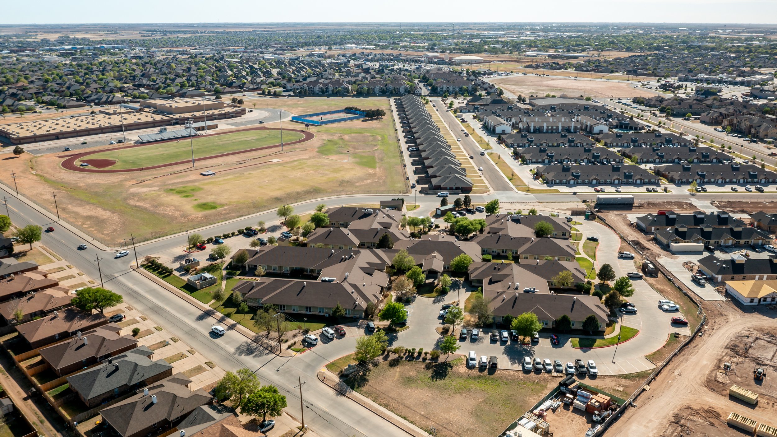 Aerial view of a senior community with residential houses, apartment complex, a running track and sports field, parking lots, and construction areas.
