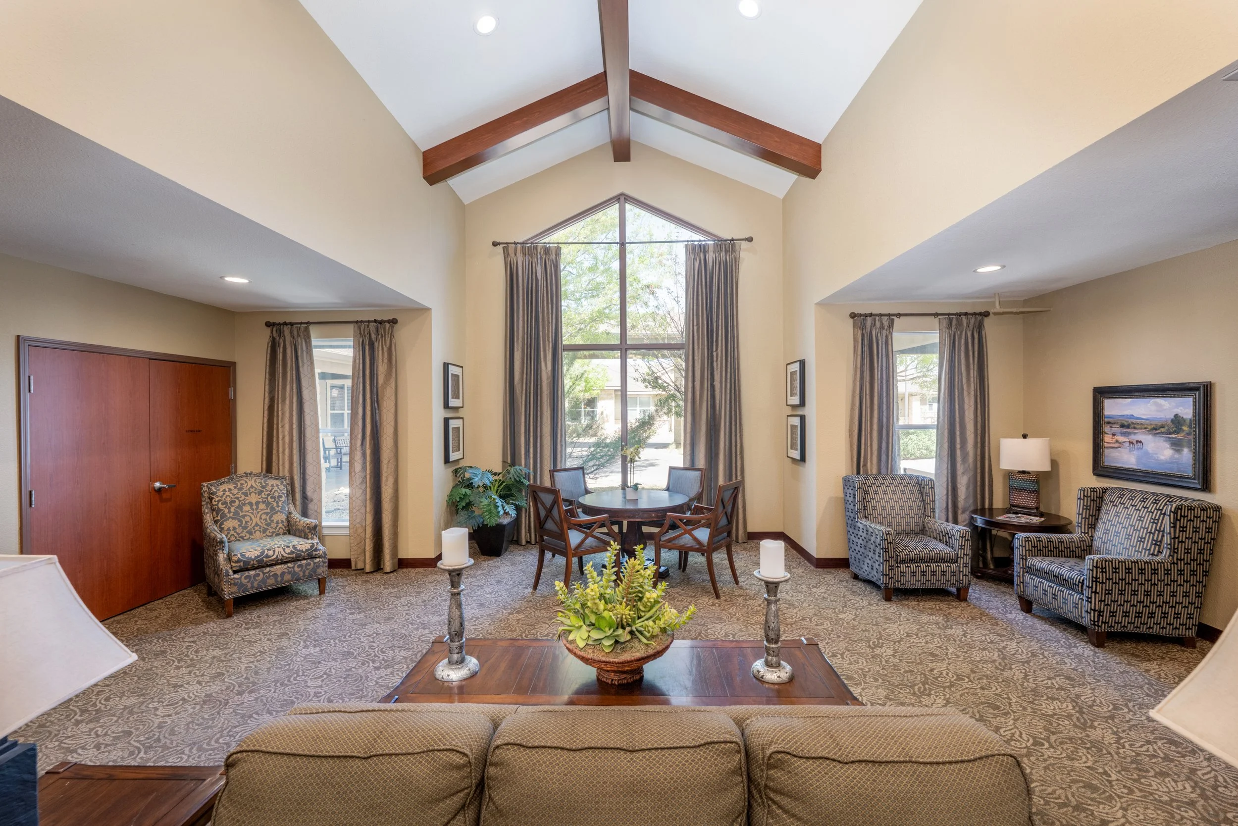 Spacious living room with high vaulted ceiling, large central window with curtains, and various seating options including a sofa, armchairs, and a small round dining table with chairs, decorated with plants, candles, and artwork.