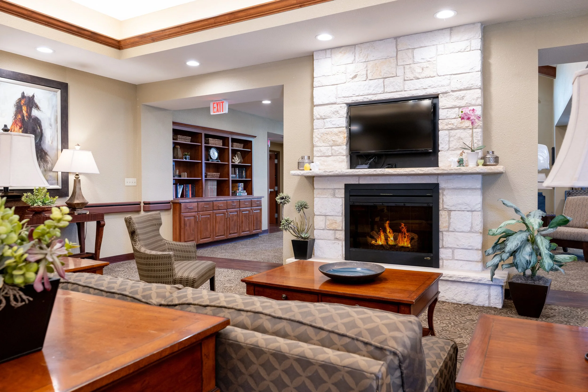 Living room with beige walls, wooden furniture, a stone fireplace with a TV above, a couch, armchairs, plants, and decor.