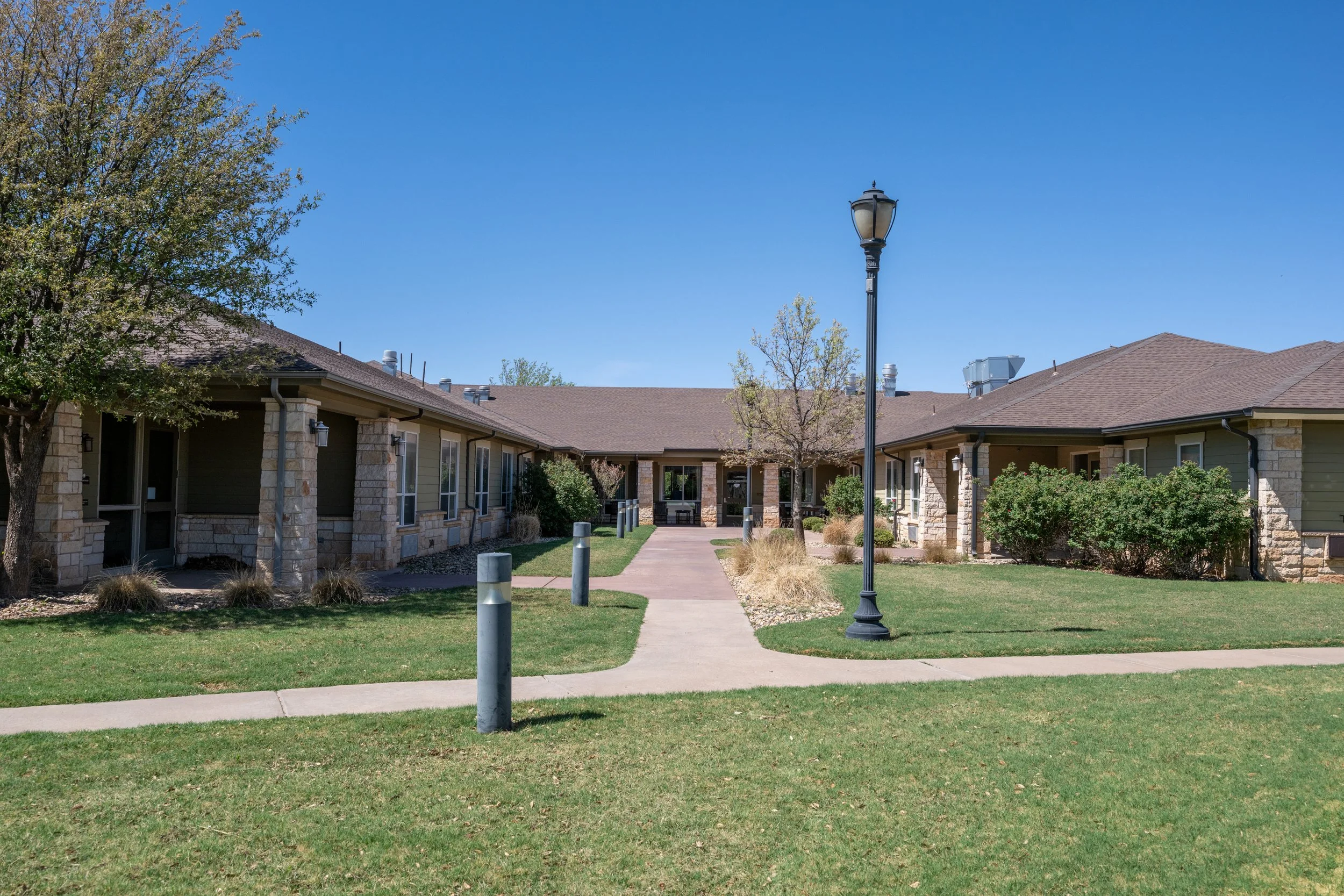 A modern nursing home with single-story buildings, landscaped yards, a paved walkway, and a street lamp under a clear blue sky.