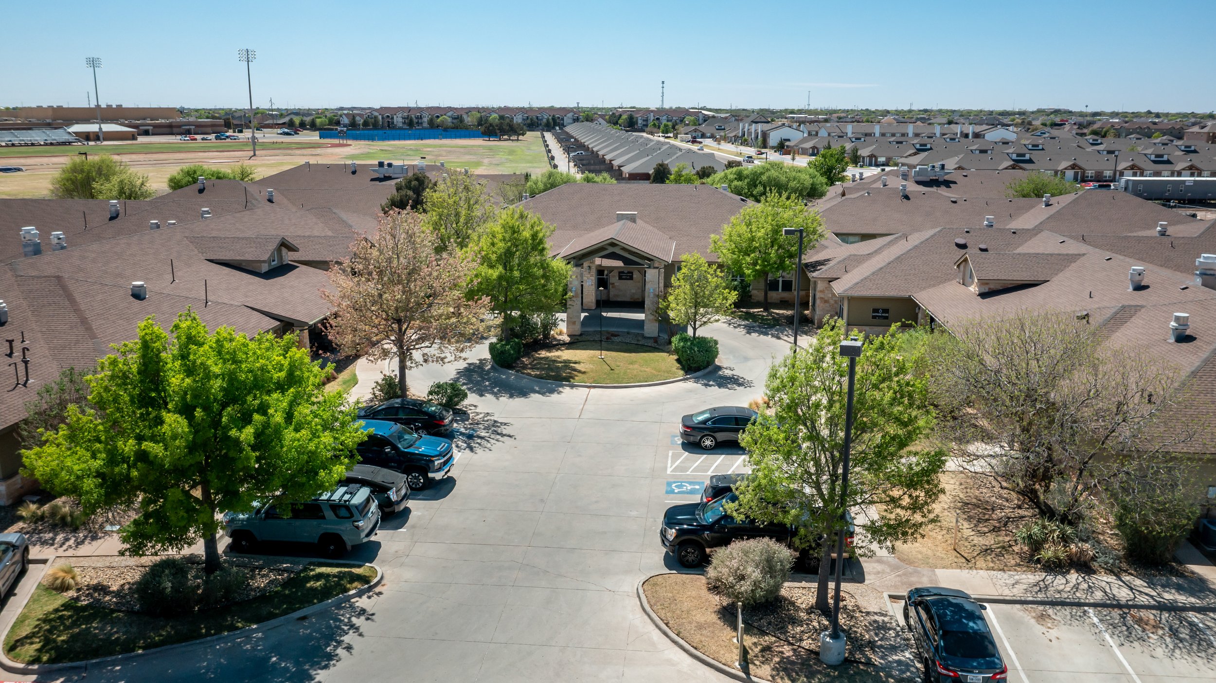 An aerial view of a post-hospital care facility in Lubbock with parking lot, trees, and surrounding houses, with sports fields in the background under a clear blue sky.