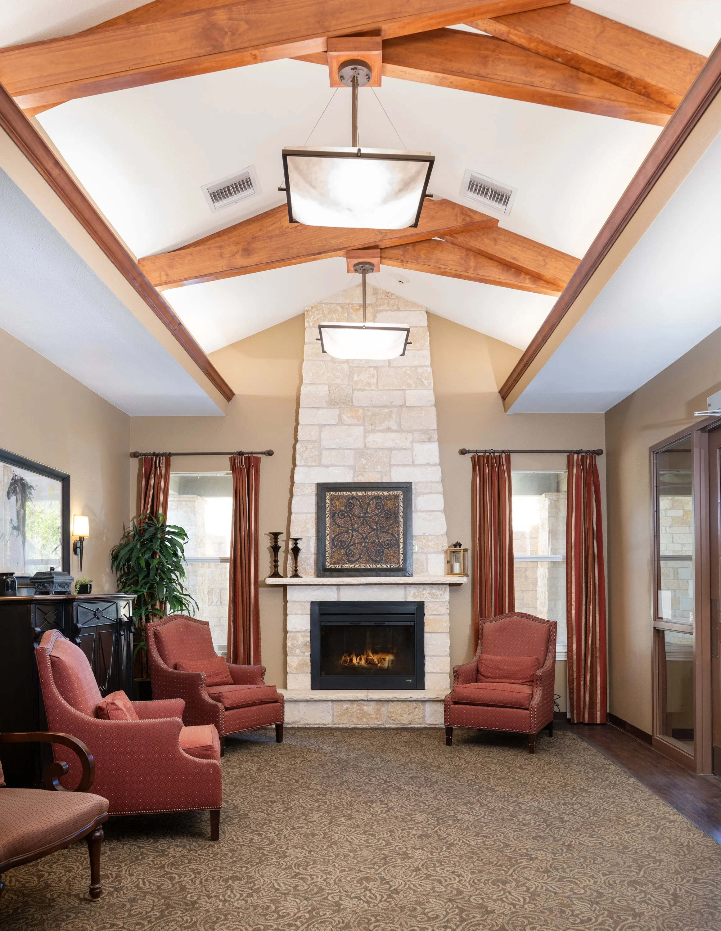 Living room with stone fireplace, wood beam ceiling, red armchairs, curtains, and a carpet.