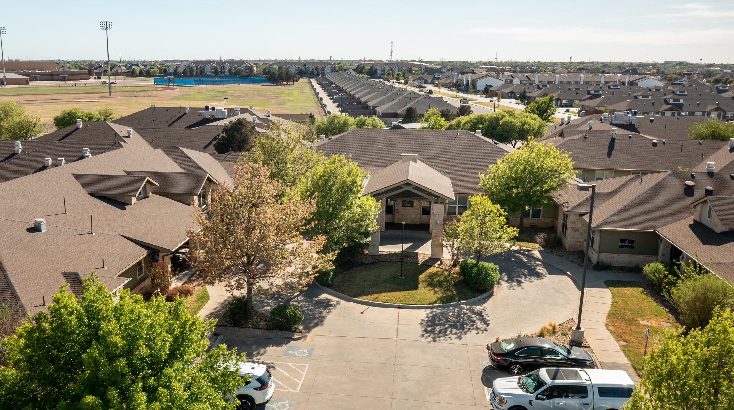 Aerial view of a senior care facility with several single-story houses, trees, and a parking lot with cars. In the background, a sports field and a row of townhouses are visible.