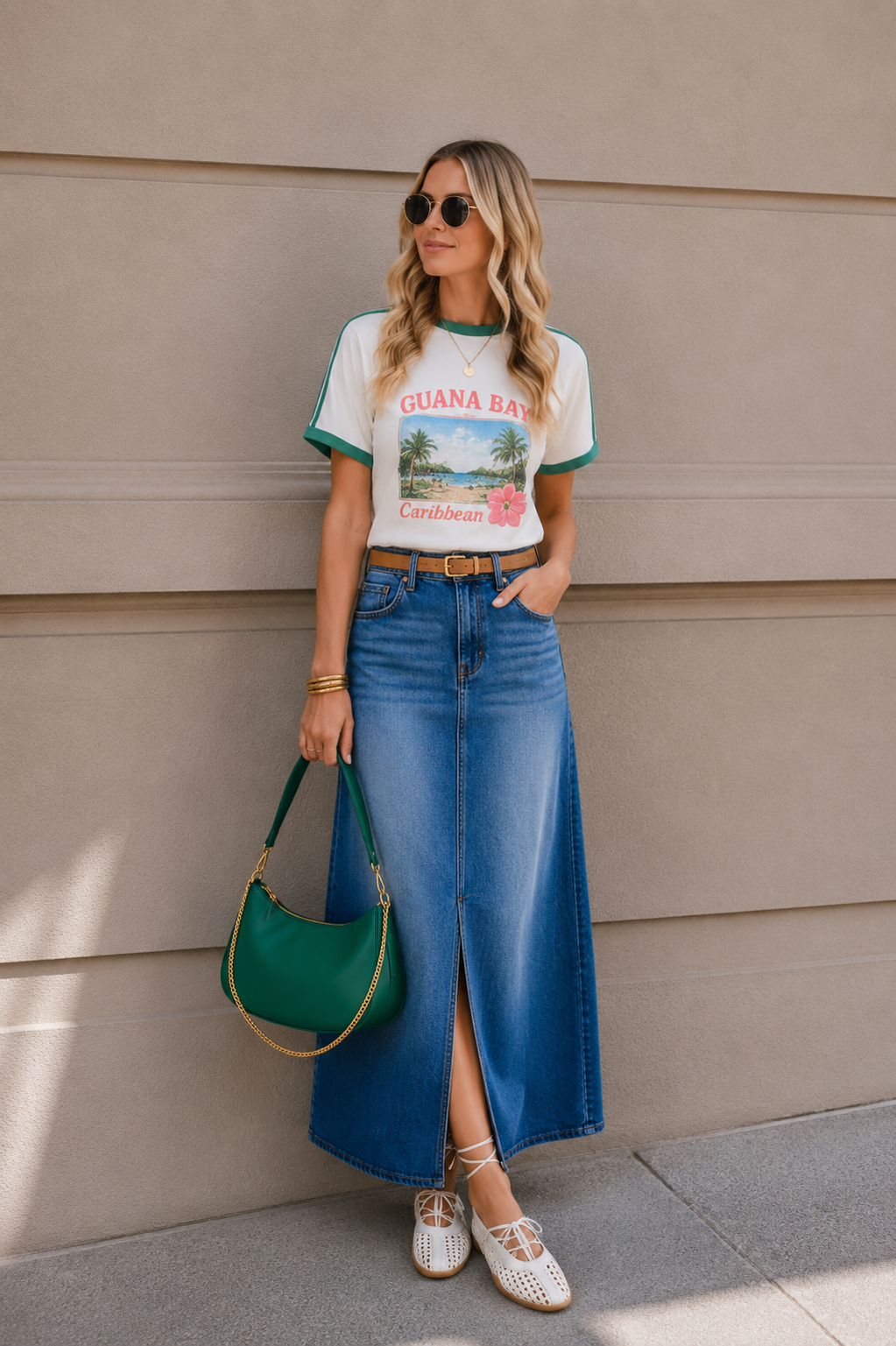 A woman standing against a beige wall, wearing sunglasses, a white t-shirt with a tropical print and the words 'Guan Bay Caribbean', a long denim skirt with a front slit, white lace-up shoes, and carrying a green handbag with gold chain details.