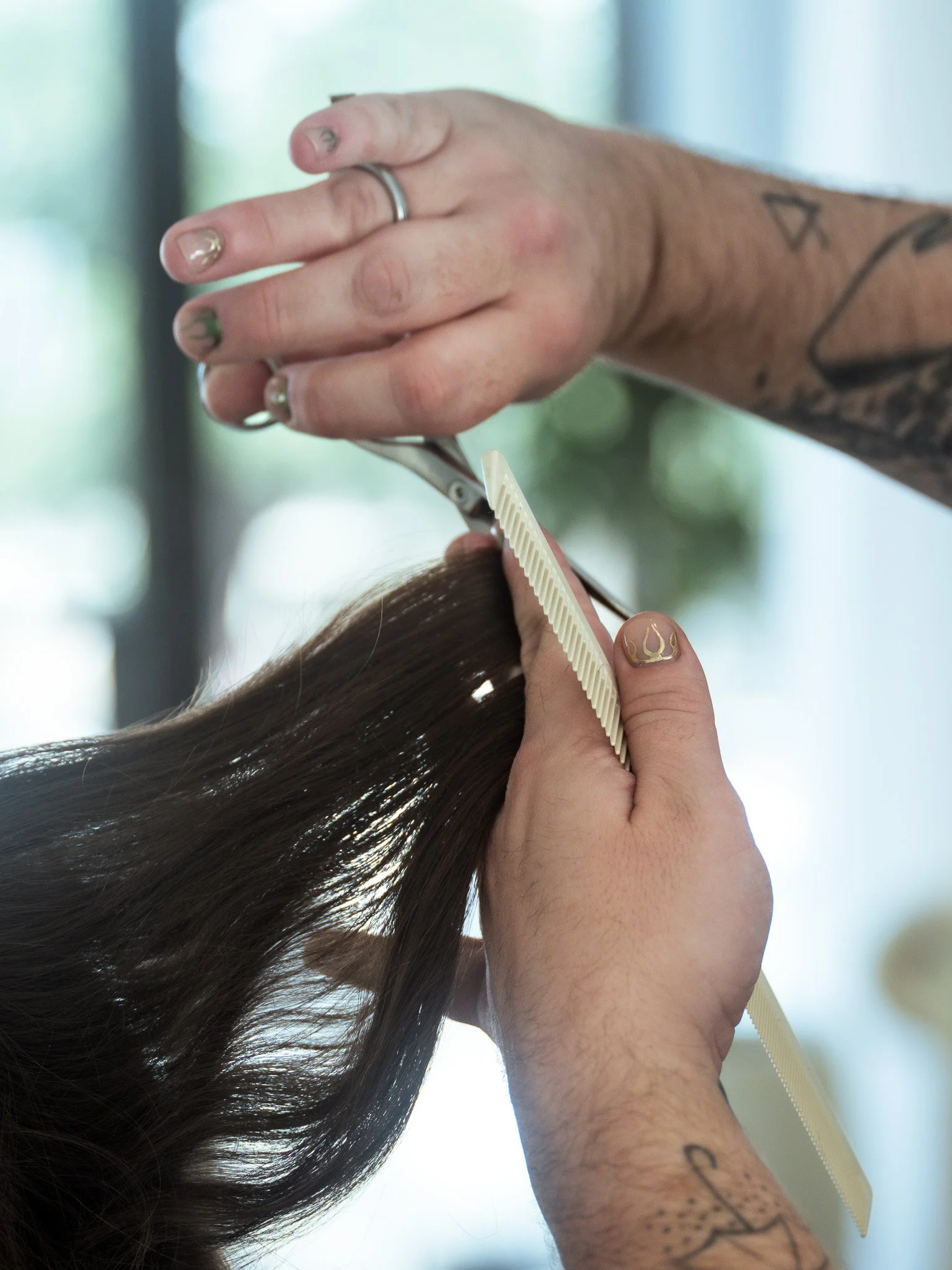 A hairstylist cuts a client's long, dark hair using scissors and a comb, with tattoos visible on their arm.