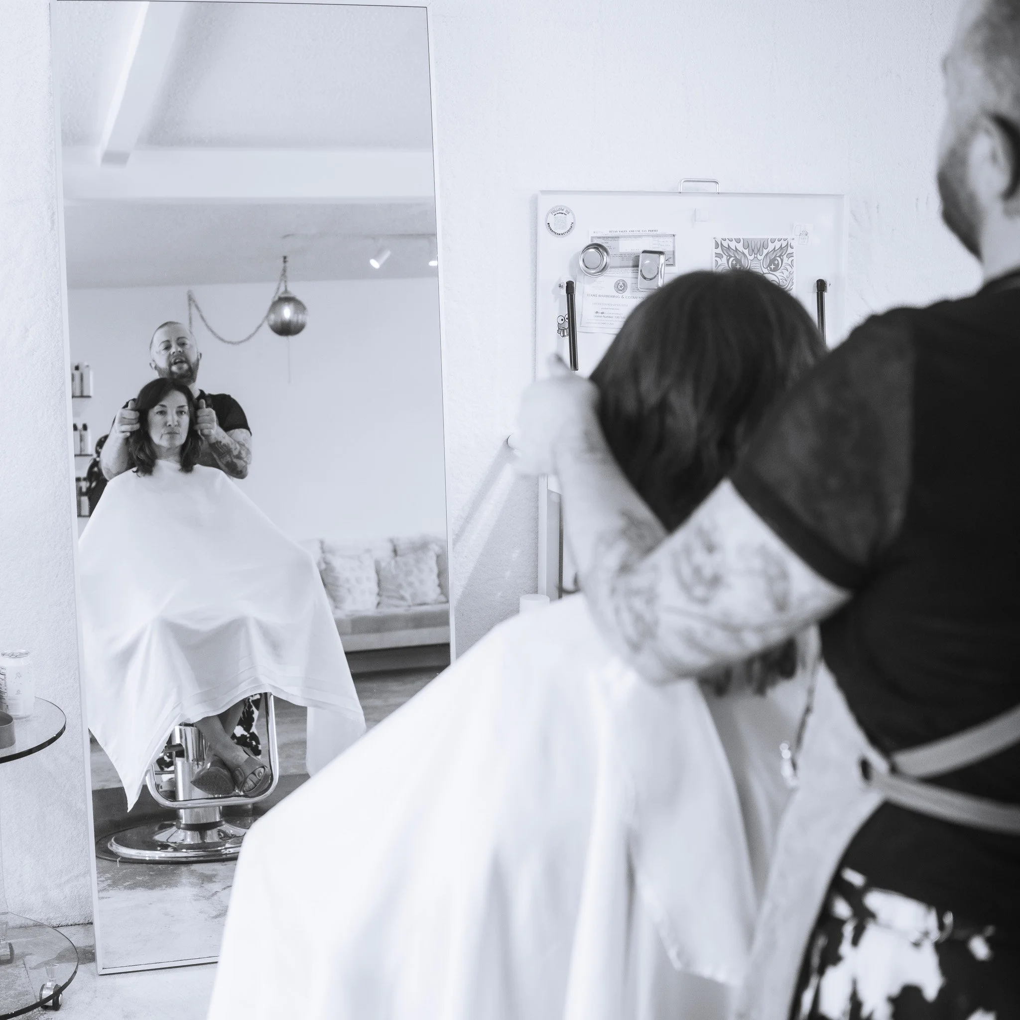 People in a barber shop looking at a mirror with a woman receiving a haircut and a barber standing behind her. A white wall is visible with a poster and a mirror reflecting part of the scene.