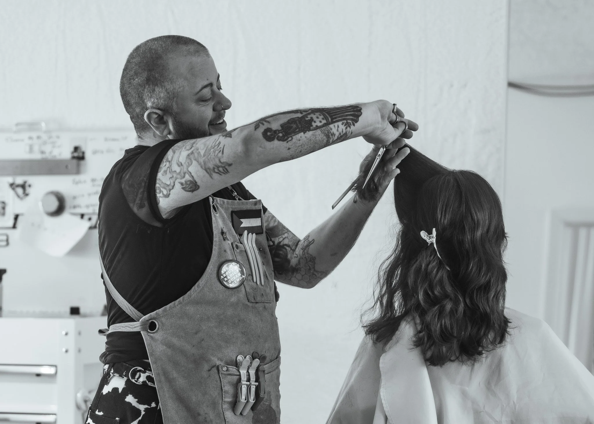 A tattooed man with a short haircut and glasses is smiling as he styles a woman's hair in a salon or similar setting.