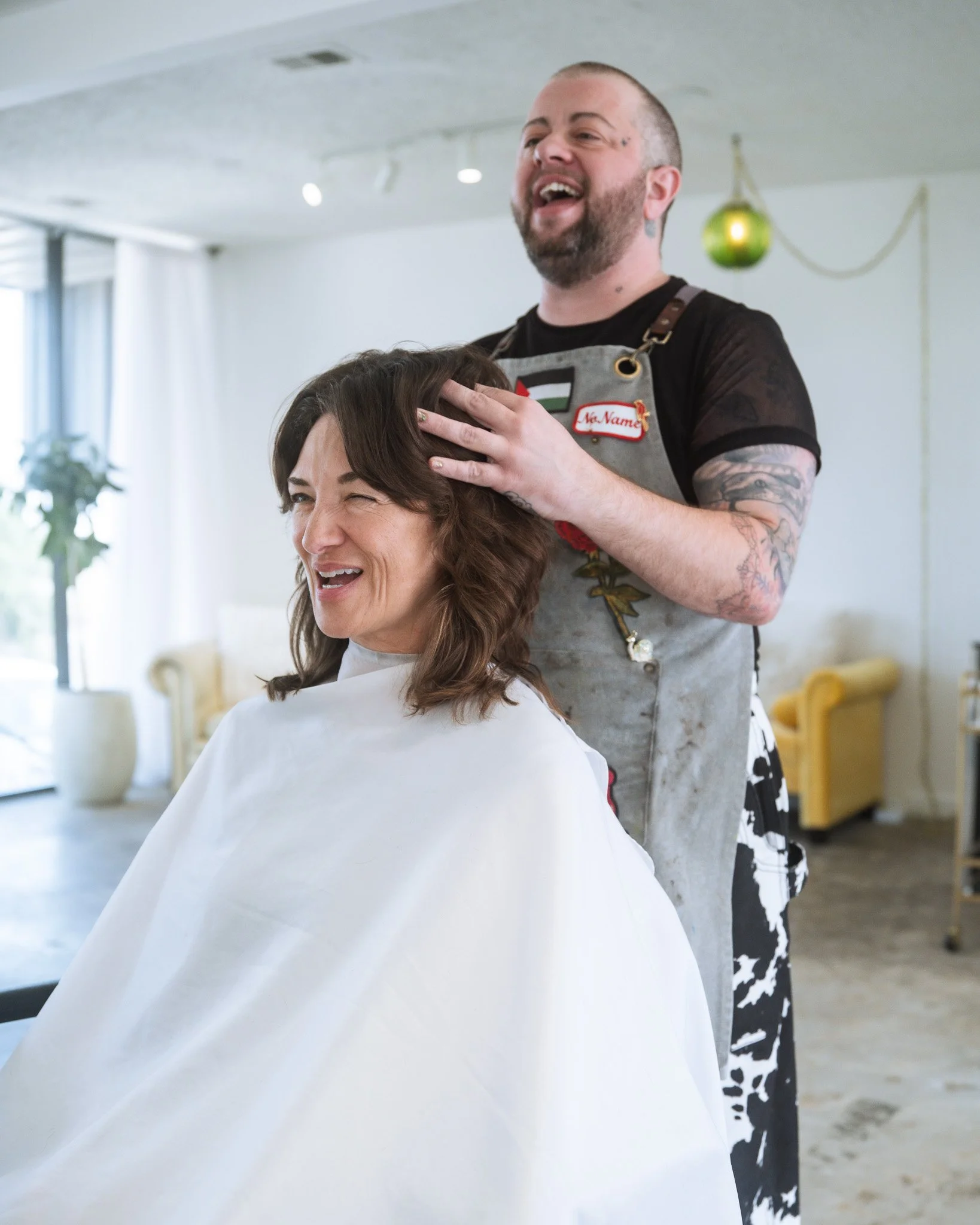 A hairstylist giving a haircut to a woman in a salon, with the stylist smiling and wearing a name tag that says 'No Name' and an apron. The salon has a bright, airy atmosphere with a hanging green light in the background.