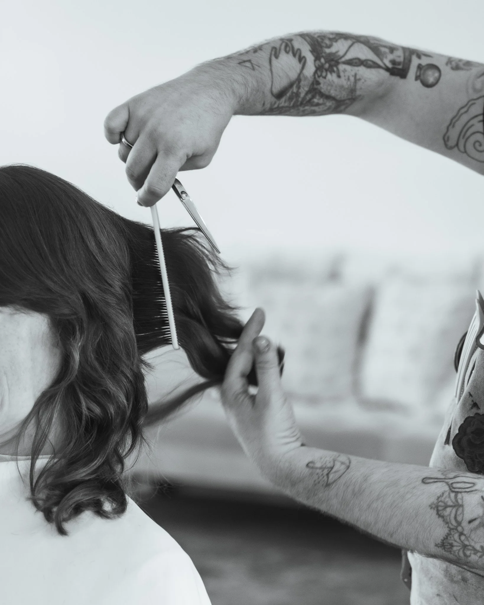 A hairstylist with tattoos on their arm is cutting a client's hair with scissors, focusing on the hair and the stylist's hand.