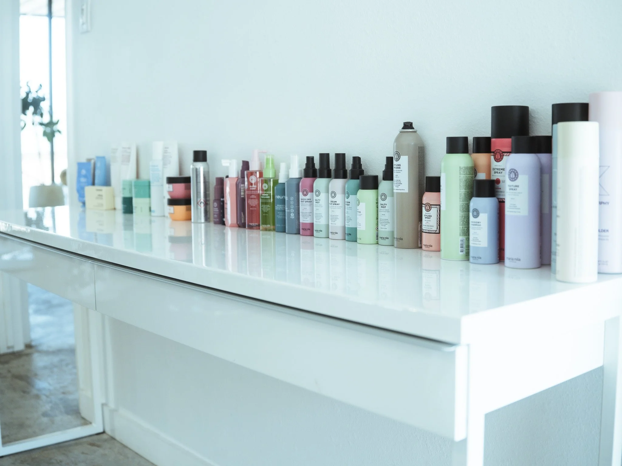 A white shelf with various hair and beauty products, including sprays, bottles, and containers, against a light-colored wall with a window to the left.