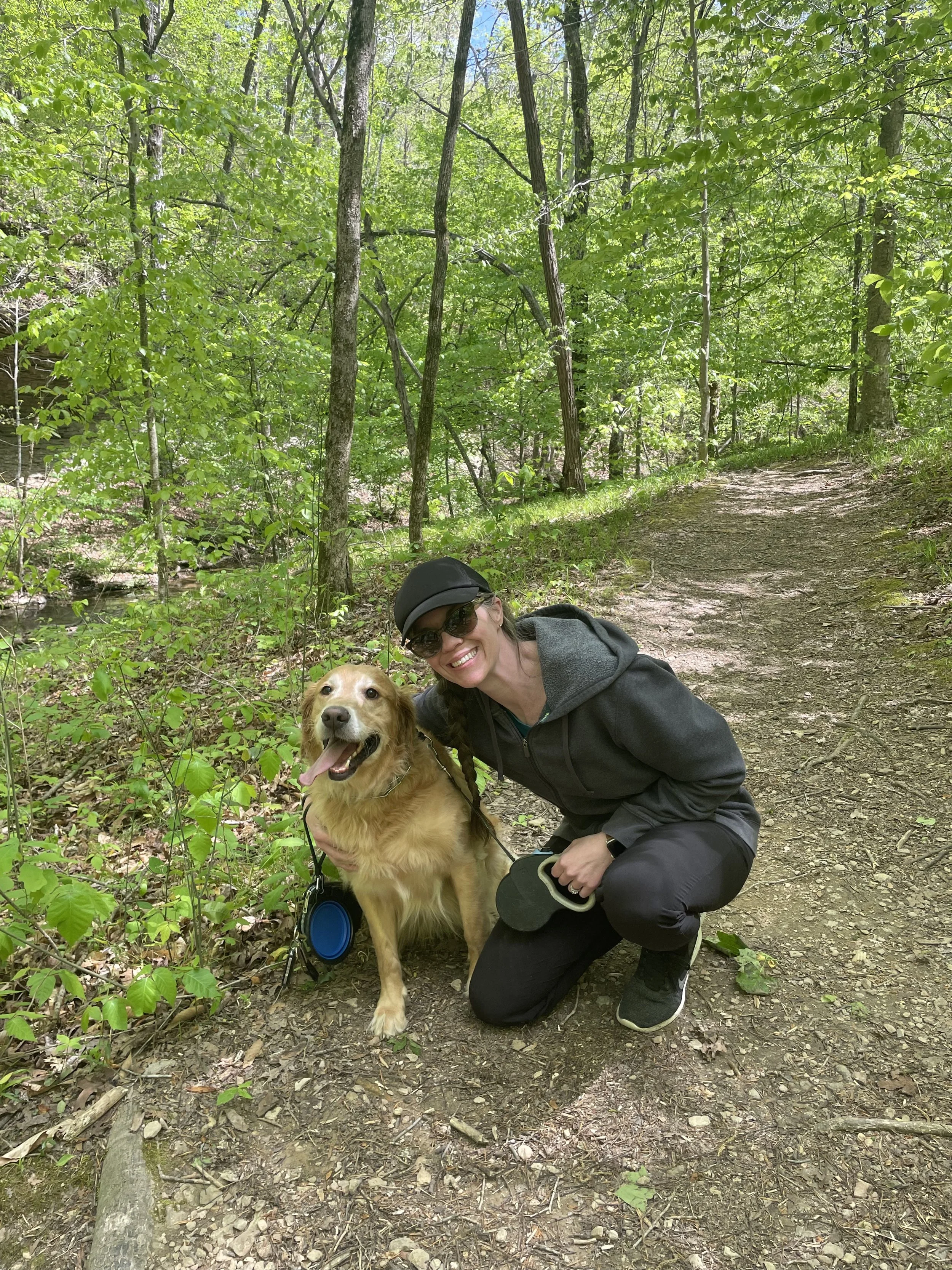 A woman smiling and crouching down next to a golden retriever on a forest trail, both appearing happy; the woman is wearing a black cap, dark sunglasses, a gray hoodie, and black pants, with a dog leash in her hand. The trail is surrounded by green trees and foliage.
