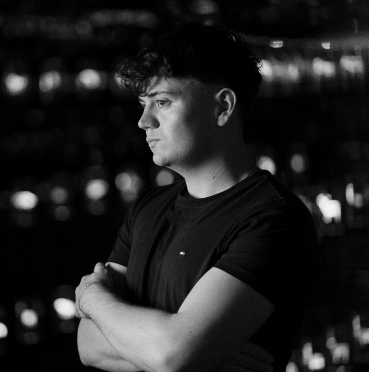 A black and white photo of a young man with curly hair, standing with crossed arms, looking to the side with a serious expression, wearing a dark T-shirt, with blurred lights in the background.