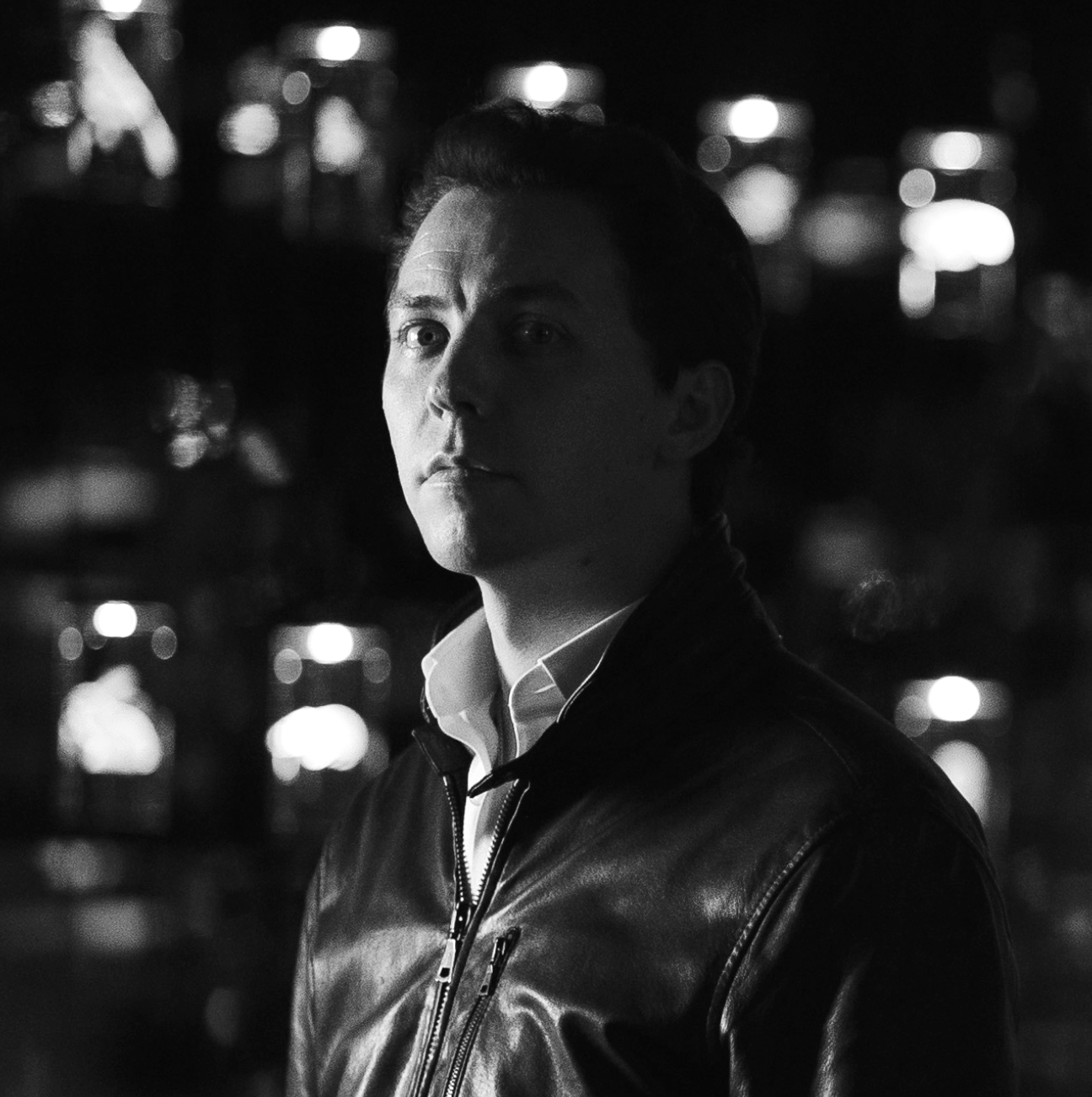 A black and white photo of a man with dark hair, wearing a leather jacket over a collared shirt, standing outdoors at night with city lights blurred in the background.