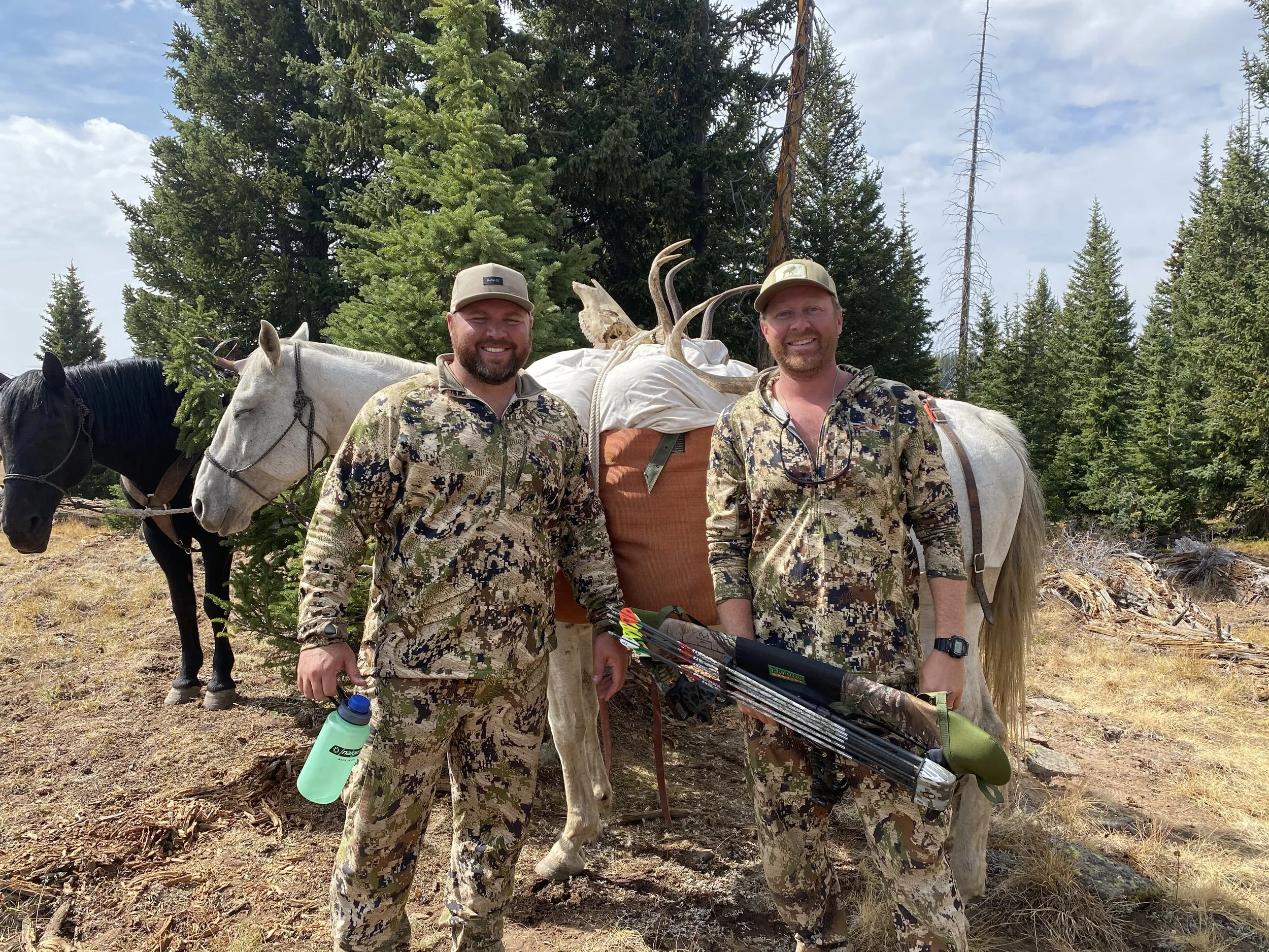 Two men in camouflage hunting outfits standing in a wooded area with a horse loaded with antlers and hunting gear, including a bow and arrows, and a green water bottle.