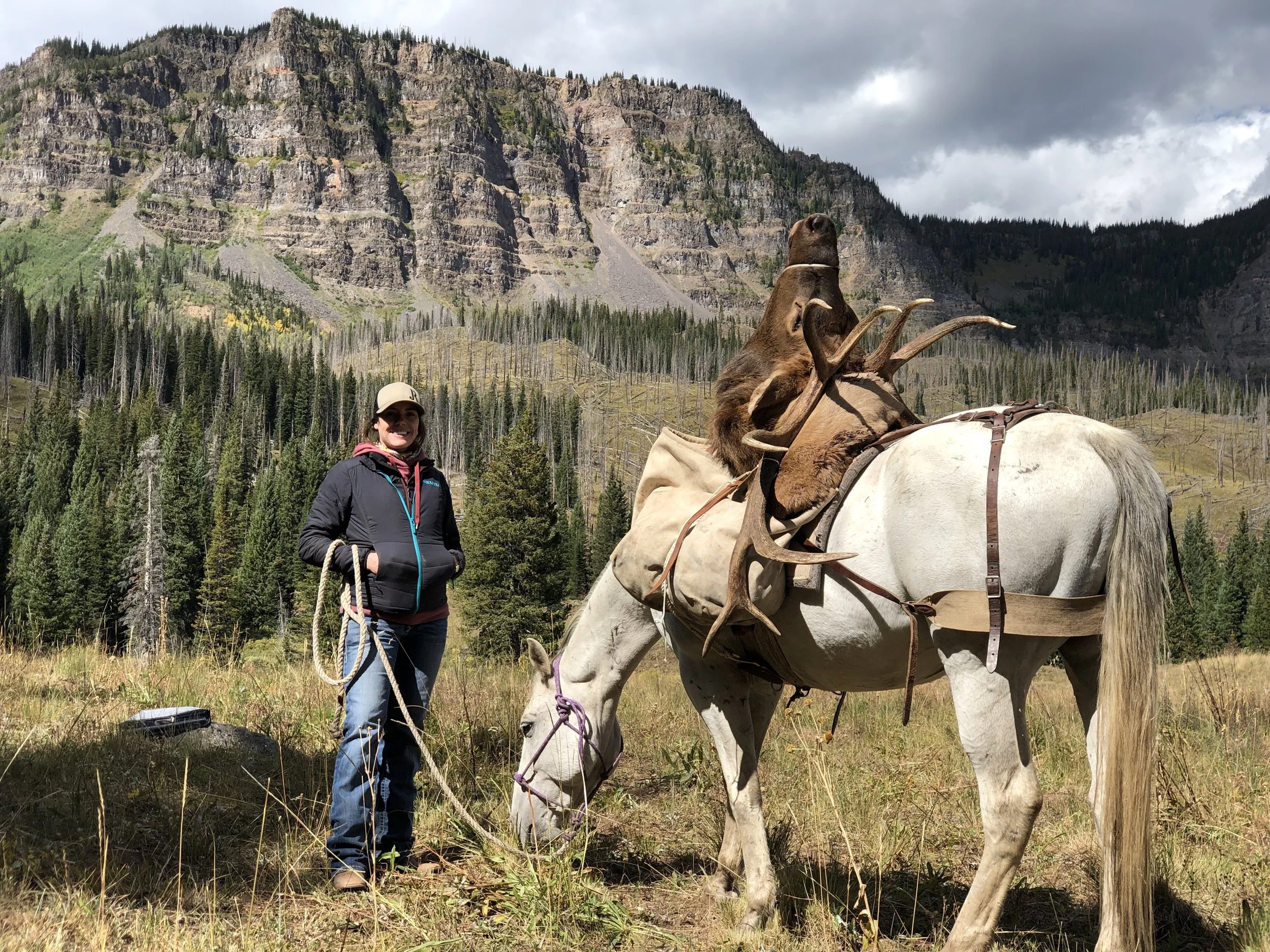 A woman standing outdoors in a mountainous forested area, holding a rope attached to a white horse with large antlers mounted on its saddle. The woman is wearing a dark jacket, a white cap, and sunglasses.