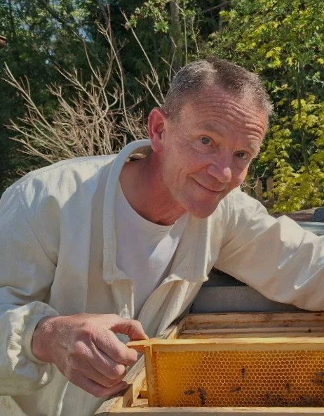 Ein Mann mit kurz geschnittenem Haar und heller Hautfarbe, trägt ein weißes Hemd und arbeitet an einem Bienenstock im Freien, umgeben von Bäumen und Pflanzen.