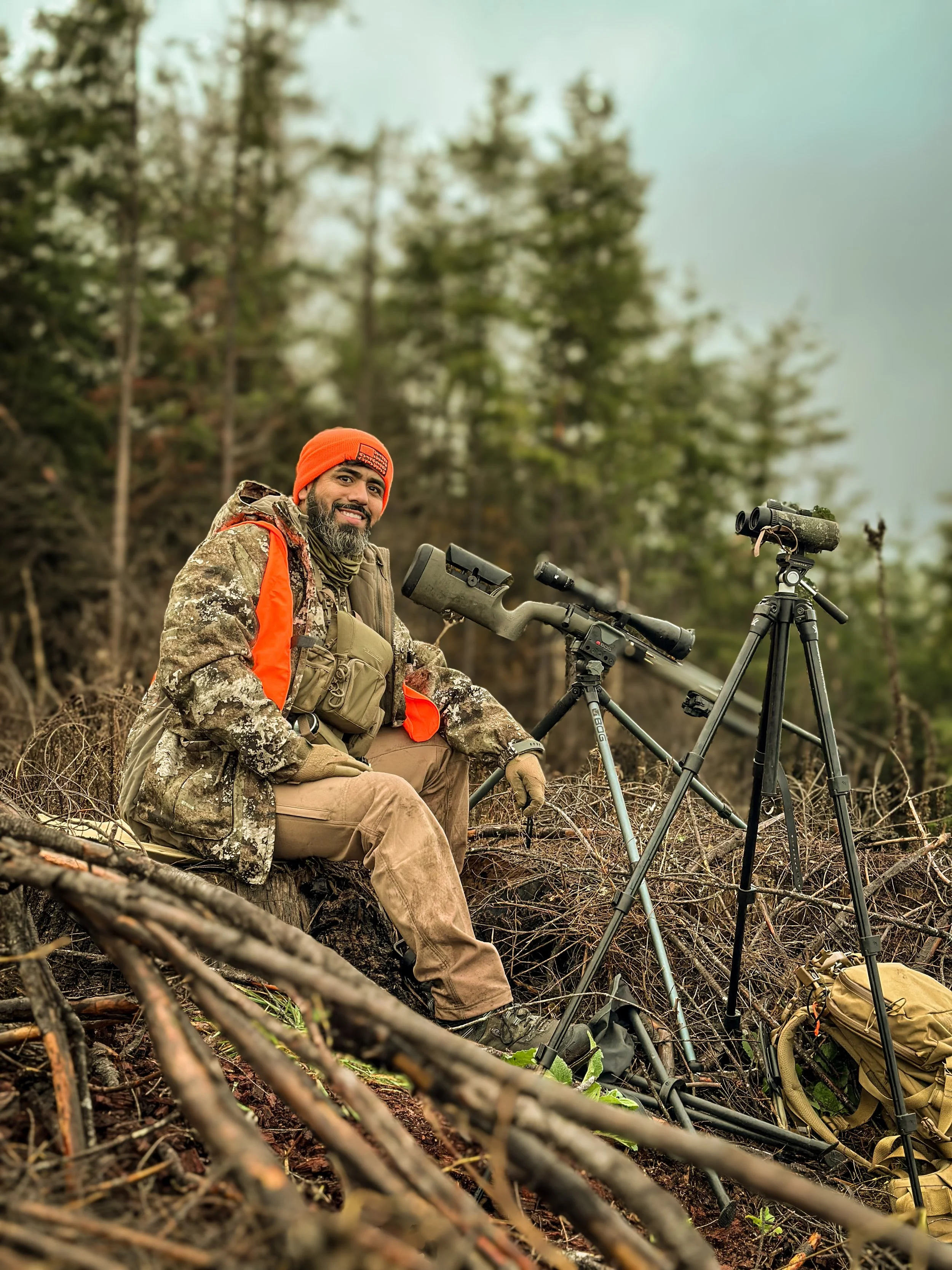 A person dressed in camouflage clothing, wearing an orange beanie, sitting outdoors in a forested area with binoculars and a spotting scope set up on tripods, preparing for wildlife observation.