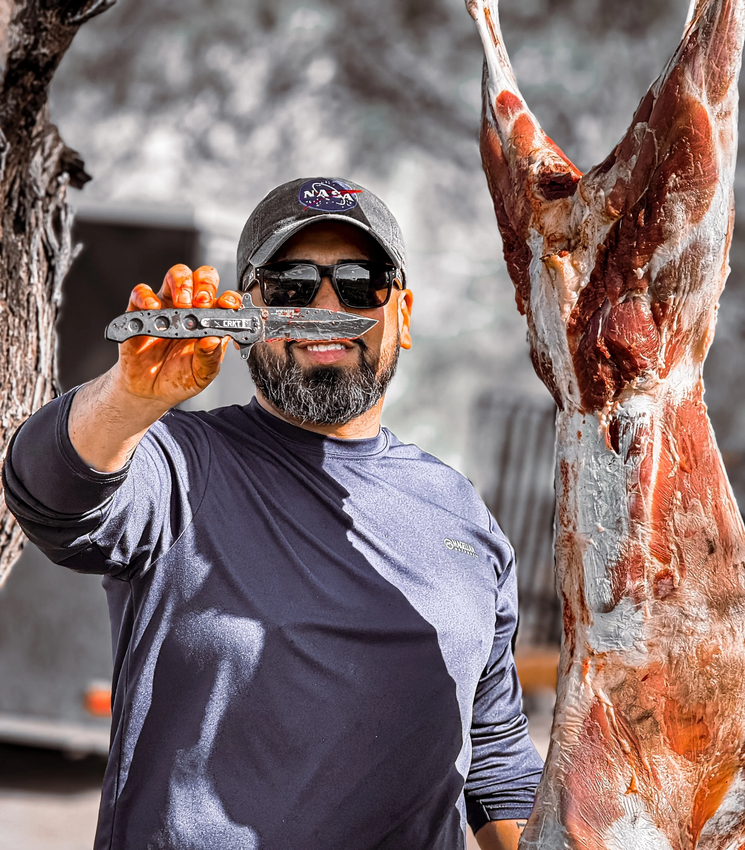 A man with a beard wearing sunglasses, a NASA hat, and a gray shirt, smiling and holding a large piece of raw meat or a carcass, outdoors with blurred trees and buildings in the background.