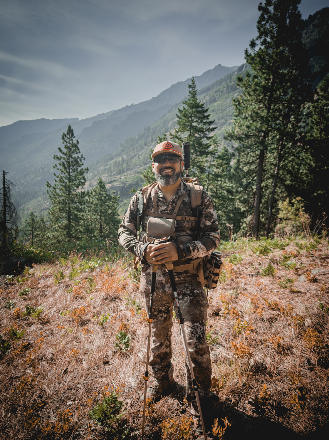 A man in camouflage clothing stands outdoors in a forested mountain area, smiling and holding hiking poles, with trees and mountain peaks in the background.