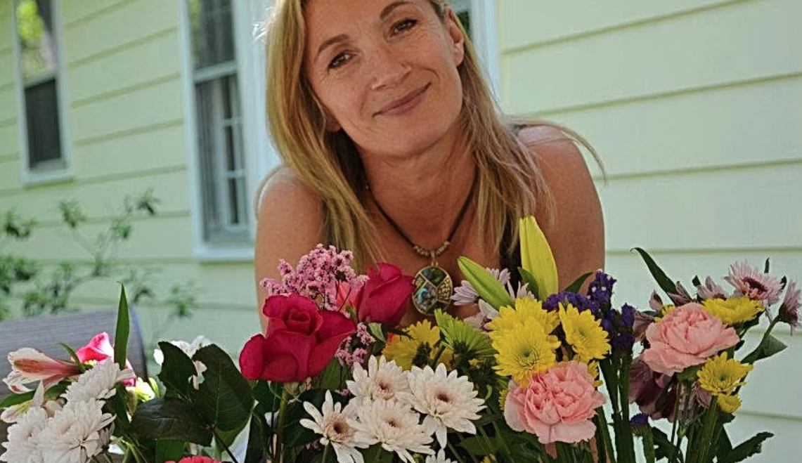 A woman with blonde hair smiling behind a colorful bouquet of flowers outside a house with light yellow siding.