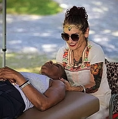 A woman with tattoos, curly hair in a bun, sunglasses, and colorful jewelry is smiling and administering a massage to an older man lying on a massage table outdoors near a body of water.