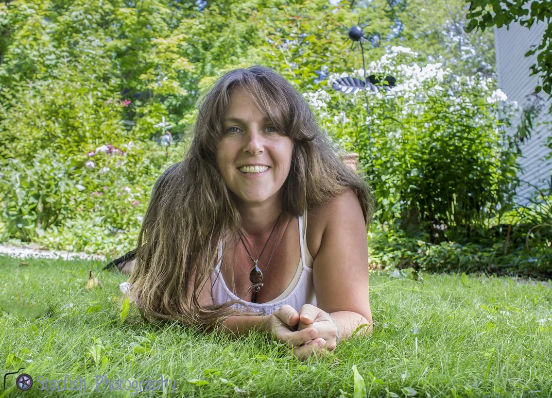 A woman with long brown hair lying on her stomach on a lush green lawn in a garden, smiling at the camera, with trees and plants in the background.