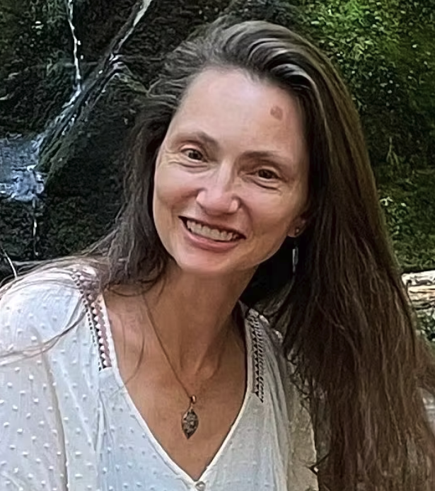 A woman with long brown hair smiling outdoors near a waterfall and green foliage.