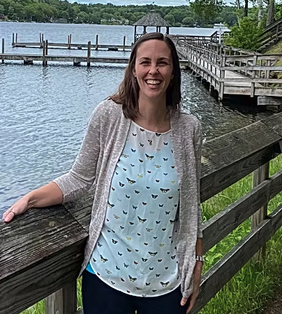A woman with long brown hair smiling, standing outdoors near a wooden fence by a lake with boats and trees in the background.