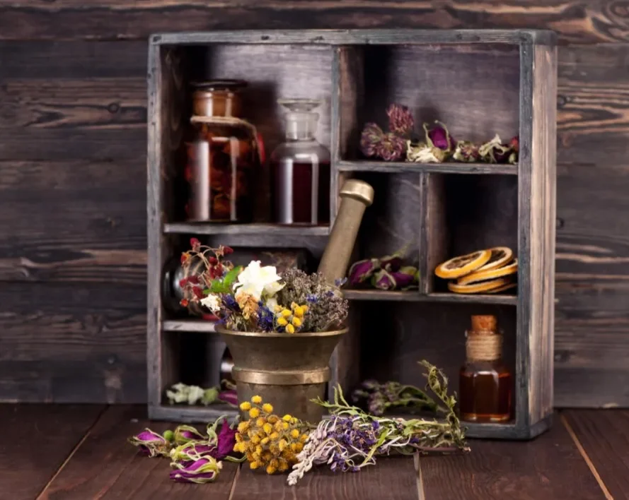 A wooden box with small compartments filled with dried herbs and flowers, some bottles of dried substances, and a mortar with fresh and dried flowers and herbs in front.
