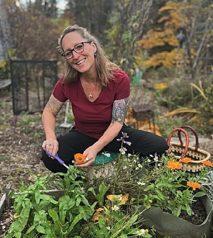 Smiling woman with glasses and tattoos in a garden, sitting on a small stool, harvesting flowers with gardening tools, surrounded by green plants and colorful flowers, autumn foliage in the background.