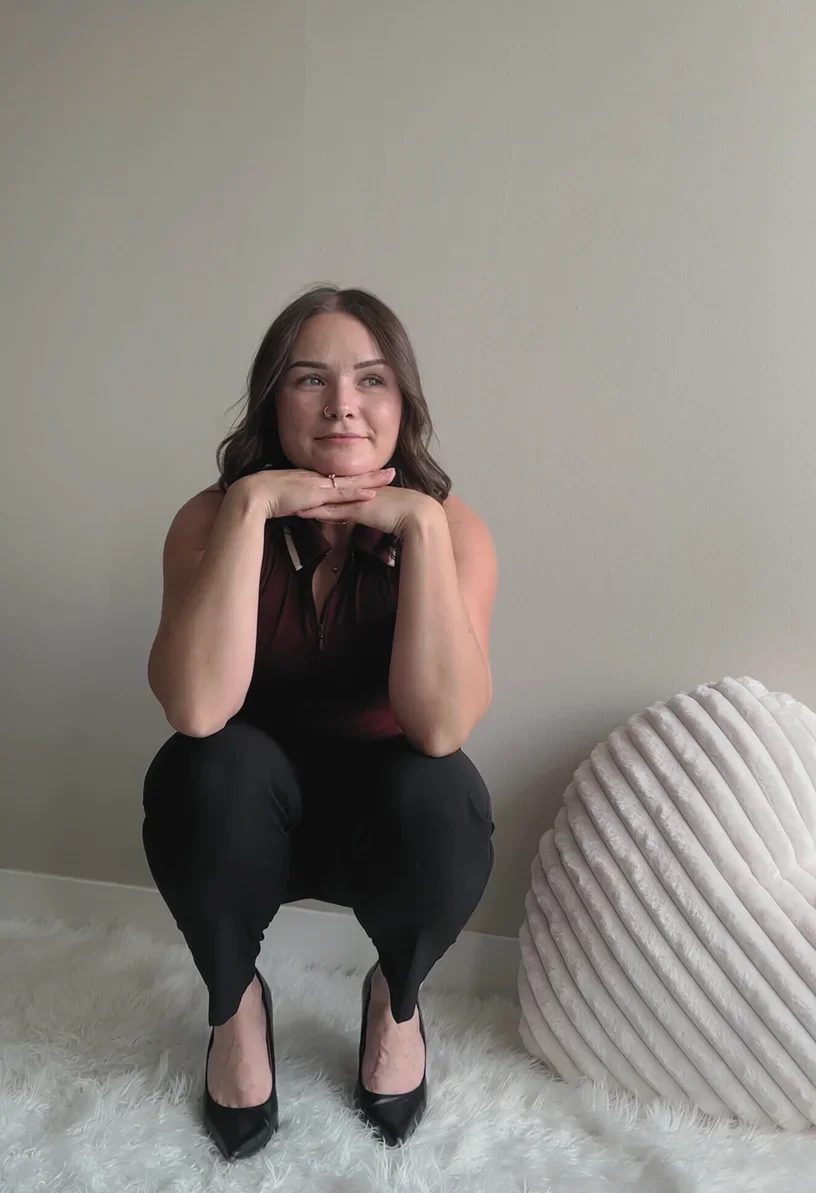 A woman squatting on a white fluffy rug against a beige wall, resting her chin on her hands, wearing a maroon sleeveless top, black pants, and black heels, with a textured round beige cushion nearby.